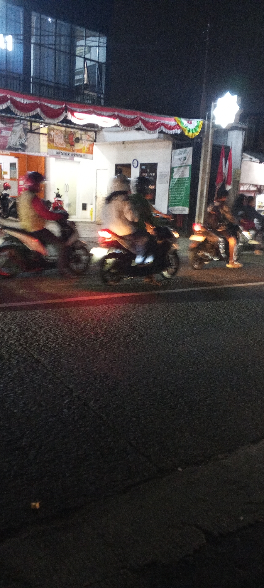 The image depicts a street scene at night in Depok, Indonesia, possibly near a pharmacy. In the foreground, a dark asphalt road shows motion blur, indicating moving vehicles. The middle ground features several motorcycles with riders, also exhibiting motion blur. One motorcycle is in the left, ridden by a person wearing a yellow vest and red sleeves. To the right of that, two more motorcycles are visible, with their riders and lights blurred. In the background, a building with large windows is illuminated, and a red and white decorative banner stretches across the top of a storefront. Signs on the building include "APOTEK ASYIFA 1" and what appears to be informational posters. A bright, star-shaped light fixture is visible on a pole to the right of the building. The overall impression is of a busy, illuminated street at night, likely in a commercial area of Depok.