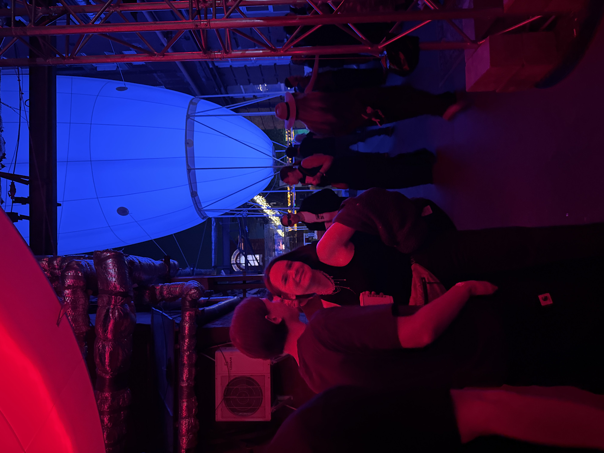 The scene, located in Moscow, Russia, depicts an indoor or semi-enclosed event space illuminated by contrasting red and blue lighting. The foreground and left side of the frame are bathed in red light, while the midground and right are predominantly blue.
In the mid-foreground, a woman with dark hair, wearing a black tank top and multiple layered necklaces, holds a white cup and smiles in the direction of the camera. To her immediate left, a person with short dark hair, clad in a dark t-shirt, stands facing towards her. A third, partially visible figure is present on the far left.
Above the left side, a large, curved red object, possibly an inflatable ceiling element, is partially visible. Beneath it, several sections of foil-wrapped industrial ductwork are horizontally mounted. A white air conditioning unit with a visible circular fan grill is affixed to a red-illuminated wall on the mid-left.
Dominating the upper midground-right is a large, blue-lit, balloon-like structure, featuring several circular apertures. To its right, a vertical red metal truss support is prominently displayed. In the background, beyond the blue structure, faint architectural details of a building facade are discernible.
Numerous other individuals are scattered throughout the midground and background, appearing indistinct due to distance and varied lighting conditions. One person on the midground-right is observed from behind, wearing a hat. The floor is dark. The environment suggests a social gathering or event within a venue featuring theatrical lighting and industrial design elements.