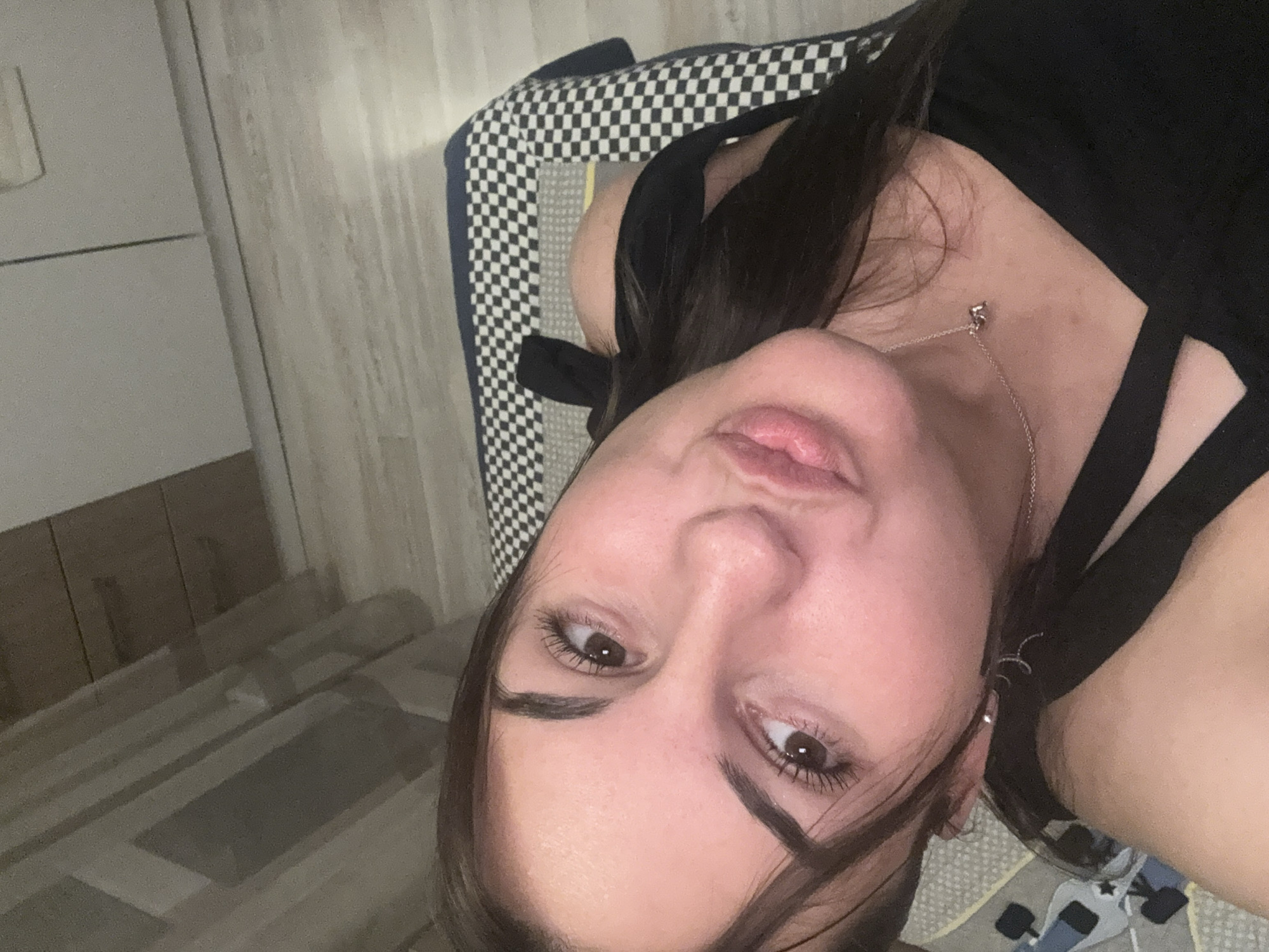 A selfie of a young, light-skinned woman. She is wearing a black top and a delicate silver necklace. Part of her dark hair falls across her face. She is lying on a surface with a black and white checkerboard pattern and a blue and gray patterned area. The background includes a beige wall and flooring. The location cannot be determined from this image.