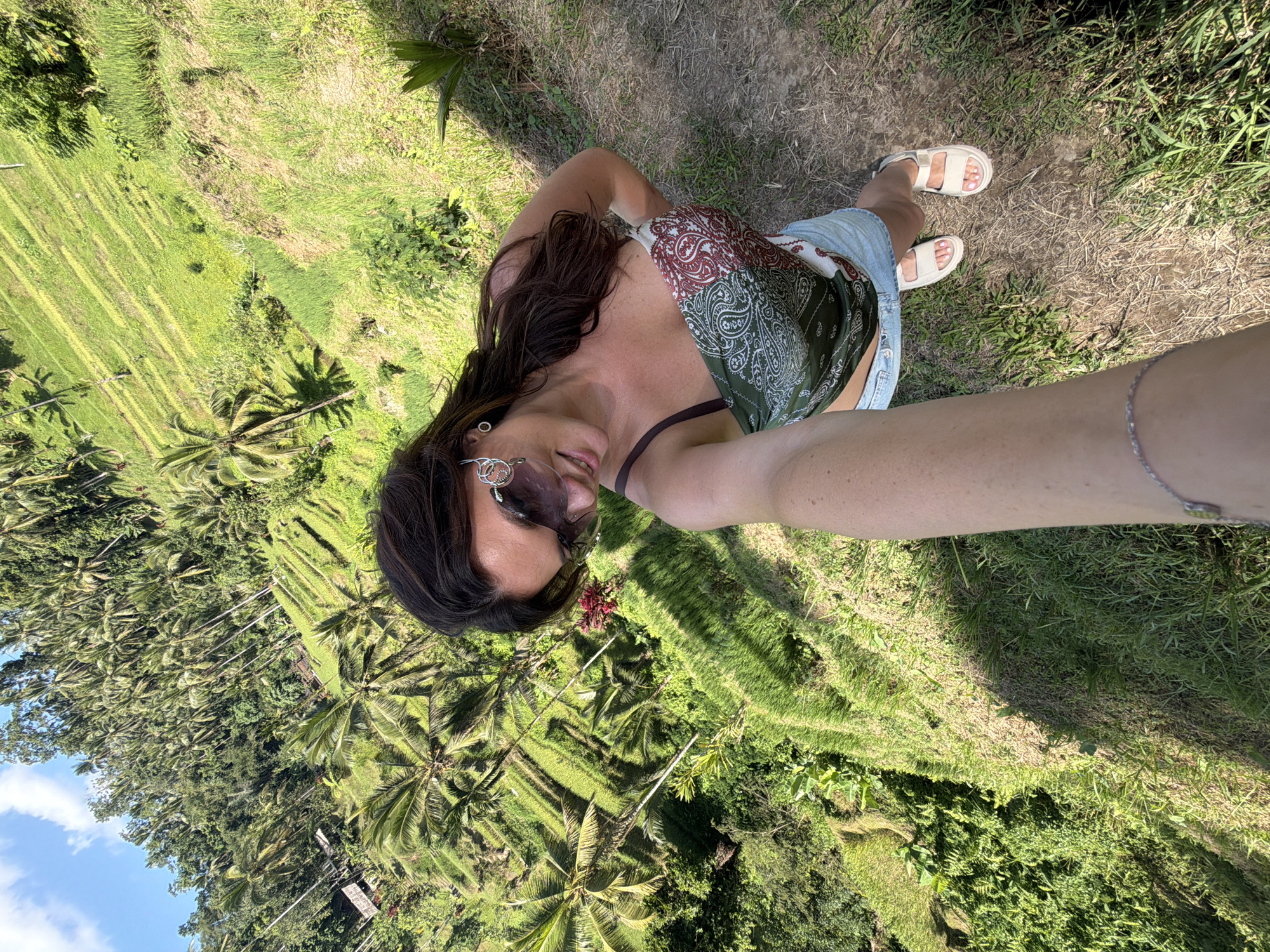 In Tegallalang, Indonesia, a woman takes a selfie from an elevated viewpoint overlooking lush, vibrant green rice terraces. The woman is positioned in the foreground, partially obscuring the view of the terraces and palm trees. She is wearing sunglasses, a patterned strapless top, and denim shorts. Her right arm, holding the camera, extends into the frame. She is wearing white platform sandals. The rice terraces descend steeply down the hillside, with some areas appearing recently harvested. The scene is bathed in sunlight, casting shadows across the landscape.