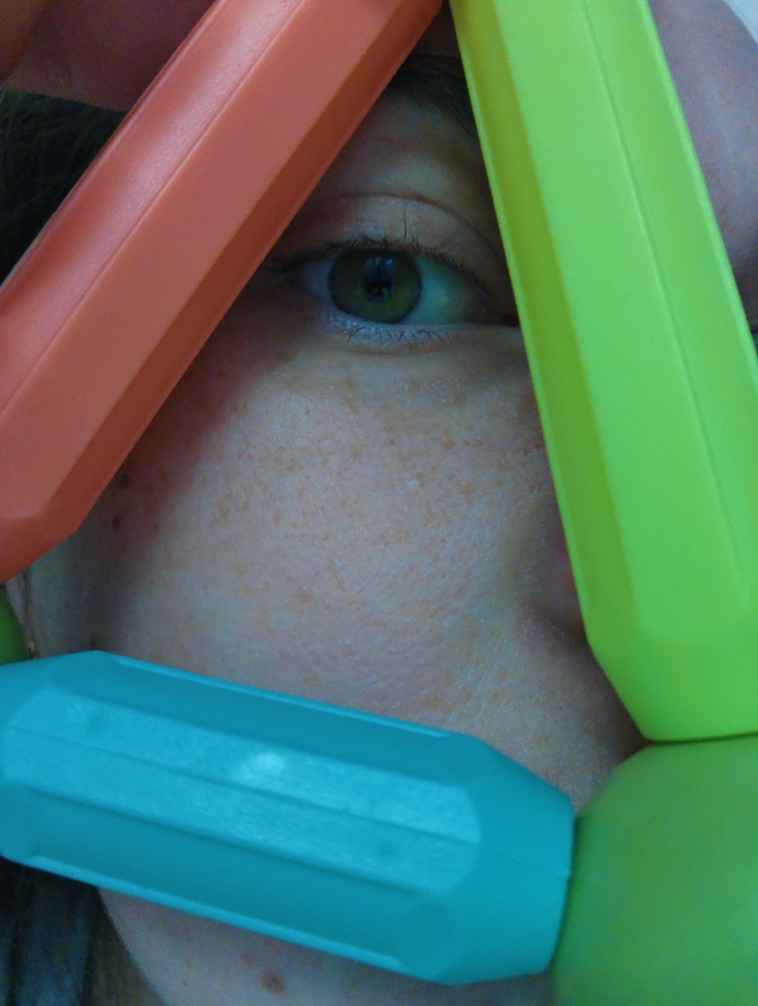 A close-up photograph captures a face partially obscured by what appear to be brightly colored, geometric plastic shapes resembling oversized construction toys. One eye is visible, exhibiting a greenish-brown iris and a natural, unenhanced appearance. The skin is fair, with freckles and subtle texture. The plastic shapes are orange, lime green, and turquoise. The face is likely indoors, illuminated by natural light, as there are no apparent artificial lighting artifacts. There is no indication of the location being Chaikovsky, Russia.