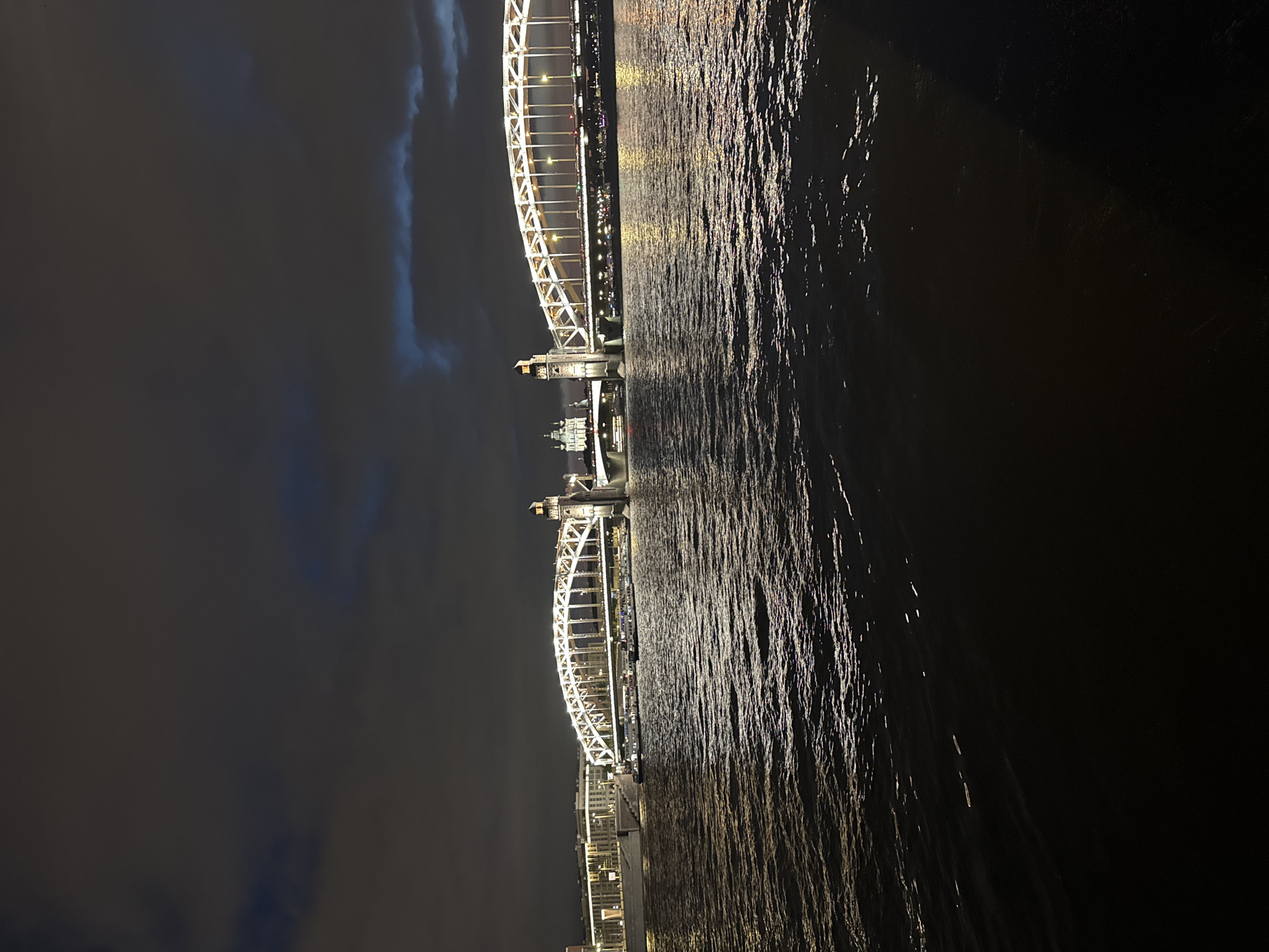 The image captures the Bolsheokhtinsky Bridge in Saint Petersburg, Russia, illuminated at night. The bridge, a prominent landmark, features an elegant arched design with white lights outlining its structure. The Neva River flows beneath the bridge, its surface reflecting the city lights and the dark, cloudy sky. Several boats are visible on the river, some docked near the embankment. The cityscape along the riverbank is dotted with buildings, also illuminated, adding to the nighttime ambiance. The sky is dark with scattered clouds, some of which are tinged with blue light, possibly from the city's illumination or residual twilight. The overall scene depicts a serene and well-lit urban landscape against a backdrop of a dim, overcast night.
