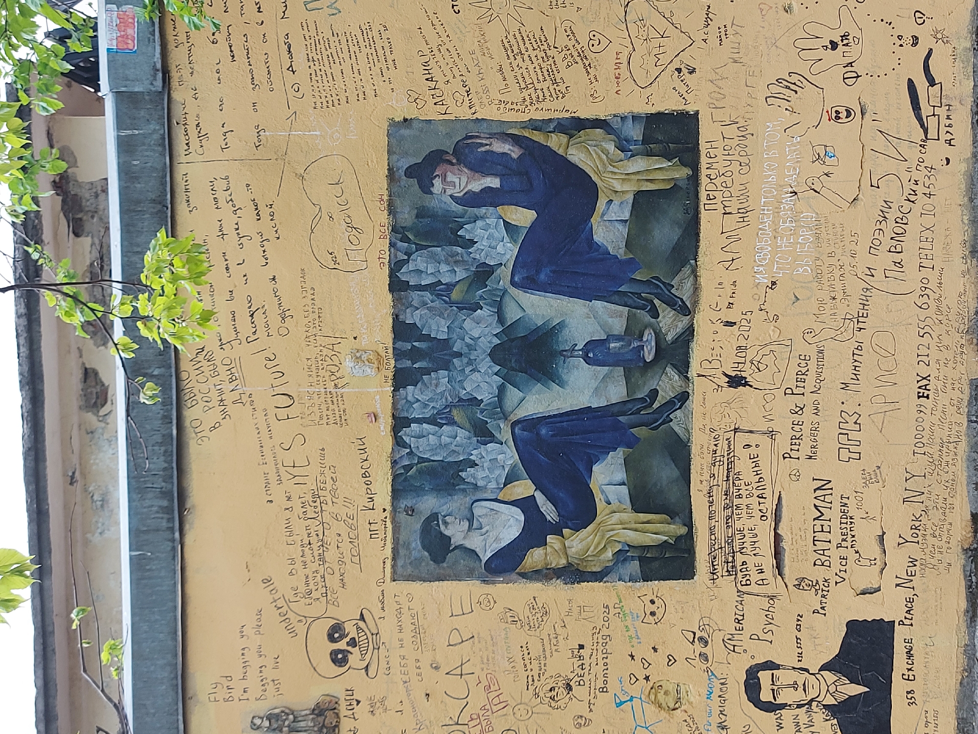 The image captures a wall in Saint Petersburg, Russia, covered in graffiti, drawings, and writings in both English and Russian. A central feature is a painted reproduction of a Cubist-style painting depicting two women. The wall's surface is yellow, and the inscriptions include names, phrases, and dates, varying in size and style. The image exhibits a mix of artistic expressions and personal messages, creating a chaotic and layered texture. Overhanging the wall, some green foliage creates a natural border at the top and left edges of the image.