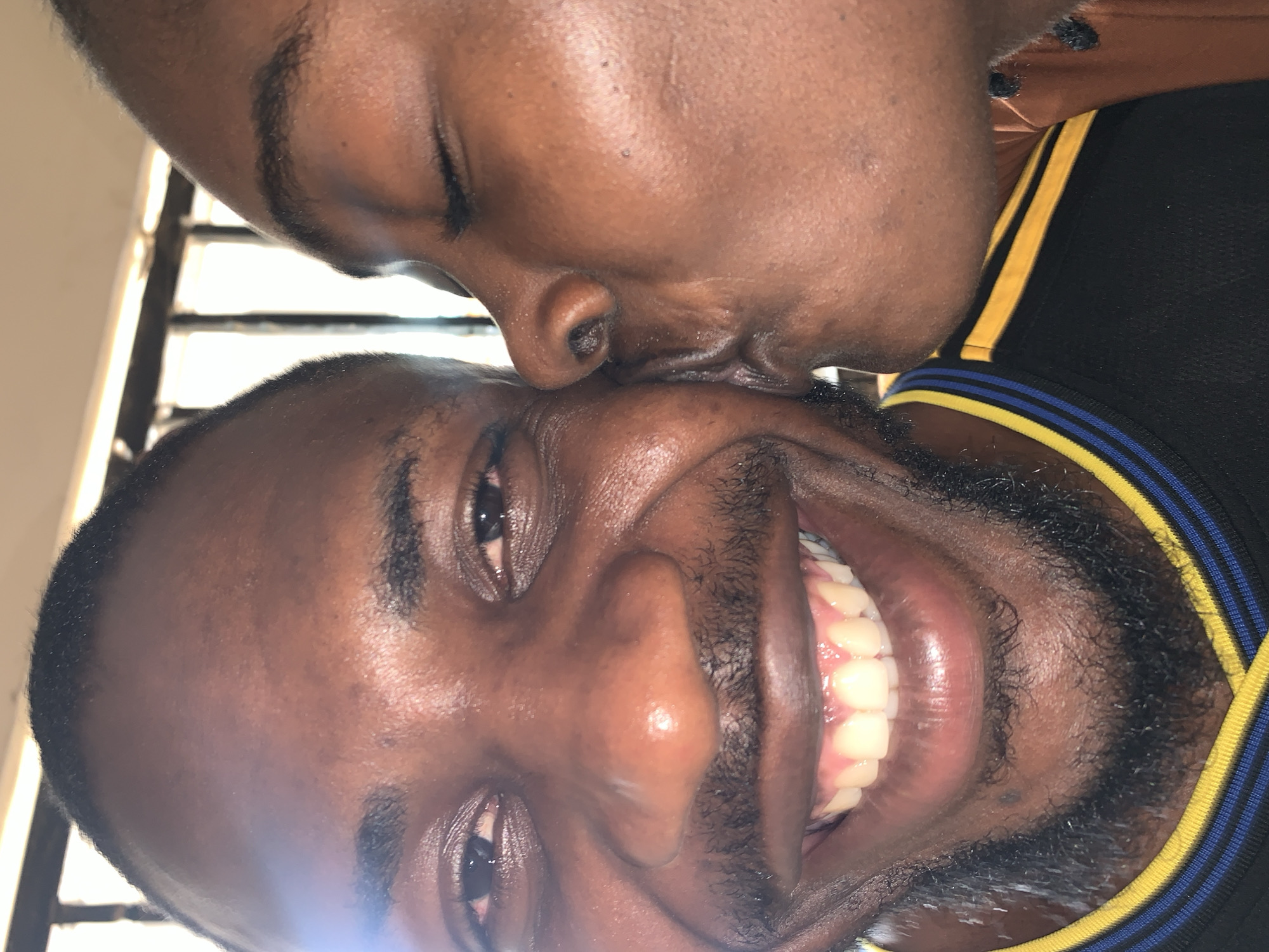 A close-up photograph taken in New Stadium, Nigeria, depicts two individuals. On the left, a person with dark skin, short dark hair, a trimmed beard, and a mustache is captured smiling broadly, displaying visible teeth and open eyes with minor scleral redness. This individual wears a black garment with yellow and blue striped trim. On the right, a second individual, also with dark skin, is shown in profile with closed eyes and pursed lips, pressing against the first individual's right cheek as if kissing. This individual's attire appears to be an orange-brown fabric. The background is out of focus, showing bright light behind dark, vertical structural elements.