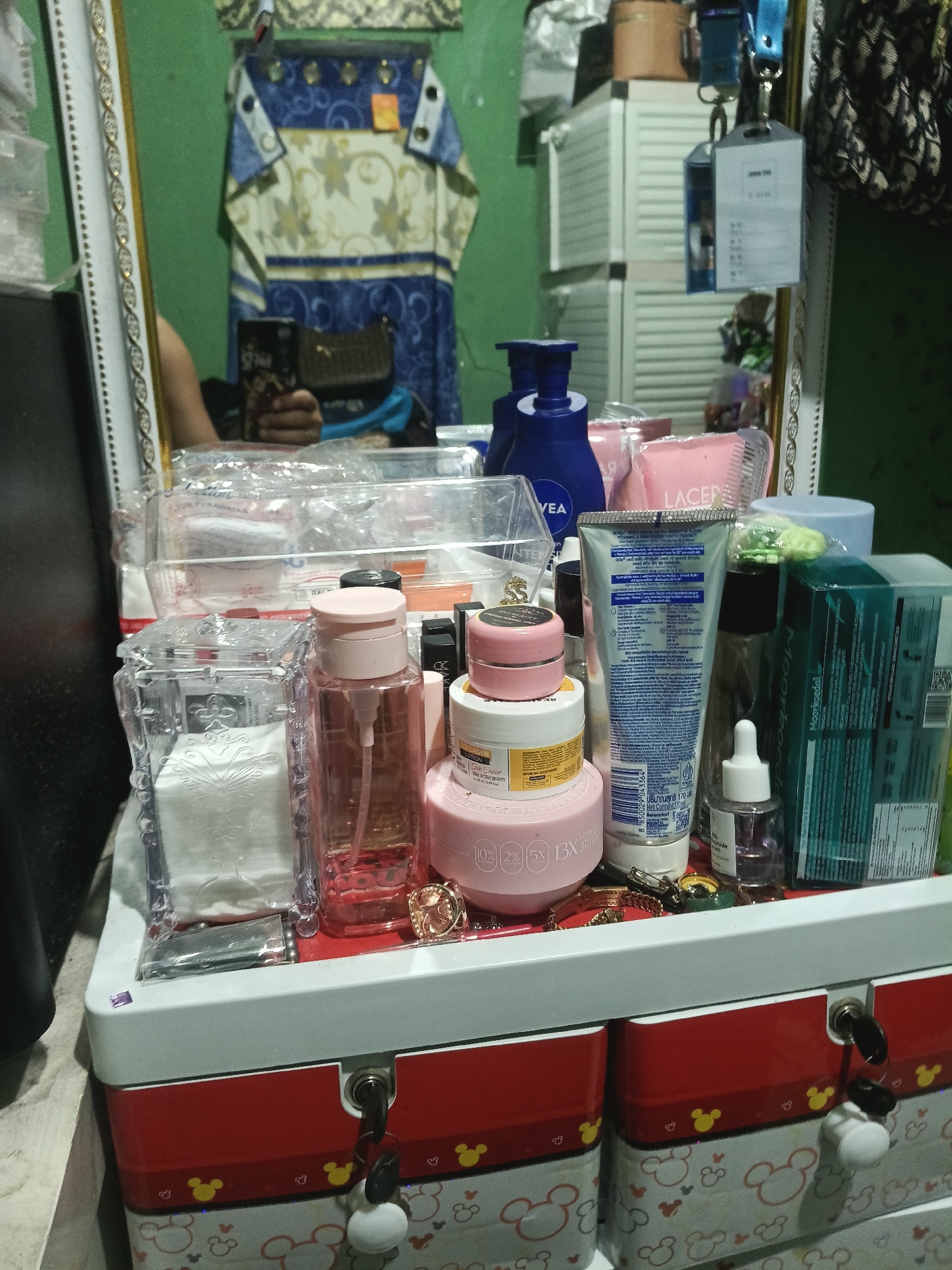 This is a close-up, eye-level photograph of a vanity or dresser top, likely in Sukamekar, Indonesia, featuring an assortment of personal care products and decorative items. The scene is dominated by various cosmetic and skincare products arranged in containers and directly on the surface. Prominently visible are several bottles and jars of creams, lotions, and toners, with labels in English and what appears to be Thai. A blue pump bottle, possibly of body lotion, stands out. Also present are cotton pads stored in a clear, decorative container, a small dropper bottle containing a liquid, and a tube of what looks like facial cream. Scattered among the products are small decorative items, including a gold-colored charm and a key. In the background, a mirror reflects a person holding a smartphone and a patterned garment hanging on the wall. The surface is a white and red multi-tiered storage box decorated with Mickey Mouse motifs, complete with small locks and keys. The overall environment suggests a personal dressing area, well-organized with beauty and personal care items.