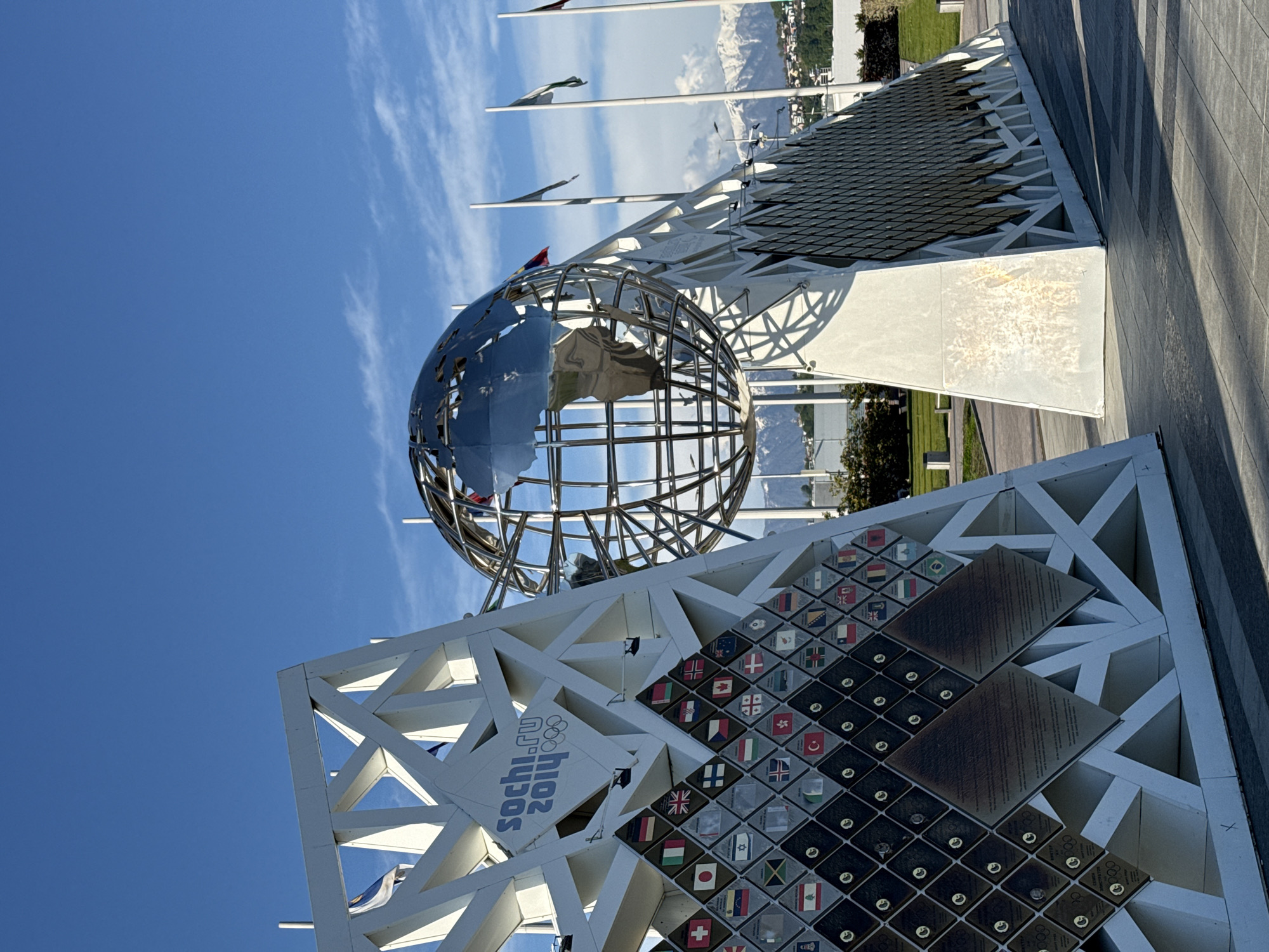 The image shows the Sochi 2014 Olympic Park in Sirius, Russia. A large metal globe sits atop a white triangular structure. The structure is decorated with the Sochi 2014 logo and plaques bearing the flags of participating countries. In the background are flagpoles, trees, and buildings, with mountains visible in the distance under a blue sky with scattered clouds. The foreground shows a paved area with linear patterns.