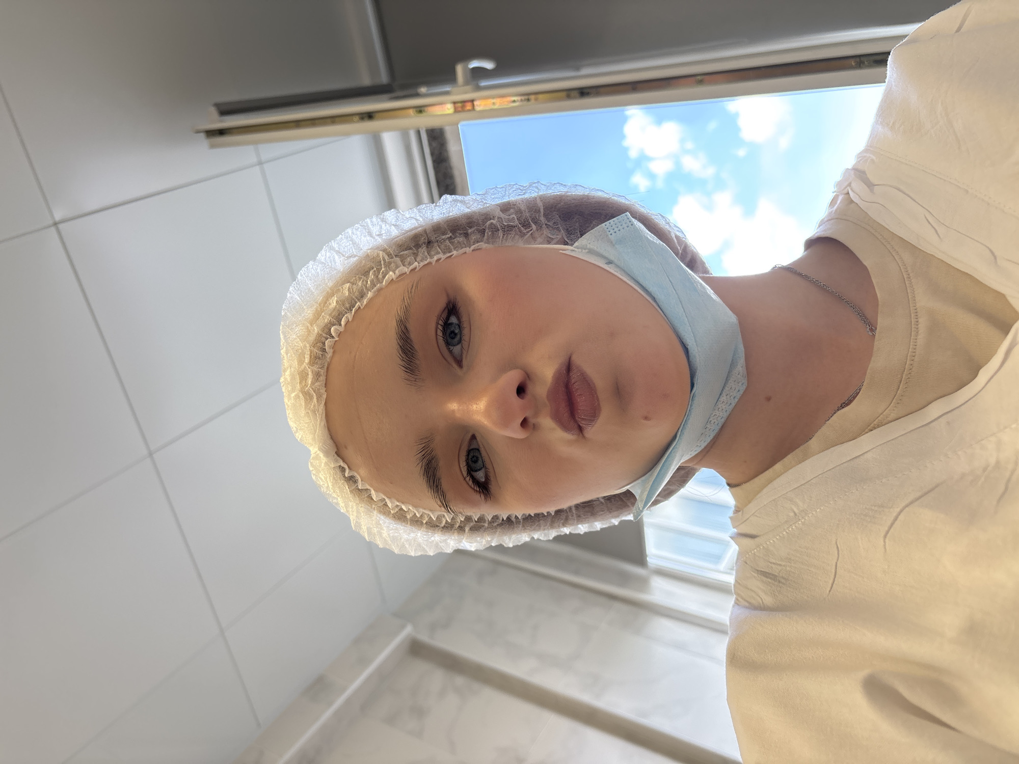 A low-angle, sideways-oriented close-up shows a young woman looking directly into the camera. She has blue eyes, groomed eyebrows, and her lips are slightly parted. Her head is covered by a white disposable bouffant cap, and a light blue surgical mask is pulled down, resting under her chin and around her neck. She wears a light-colored, possibly off-white, t-shirt, and a thin silver chain necklace is visible.
The background features white tiled walls, typical of a medical or cosmetic setting. Above her head, a window reveals a clear blue sky with scattered white clouds, suggesting daytime. A window frame and what appears to be a light fixture or strip lighting are also visible at the top. The overall scene implies an indoor environment, possibly a professional clinic or facility, contextually located in Saint Petersburg, Russia. The woman's expression is neutral and calm. There is no visible text in the image.