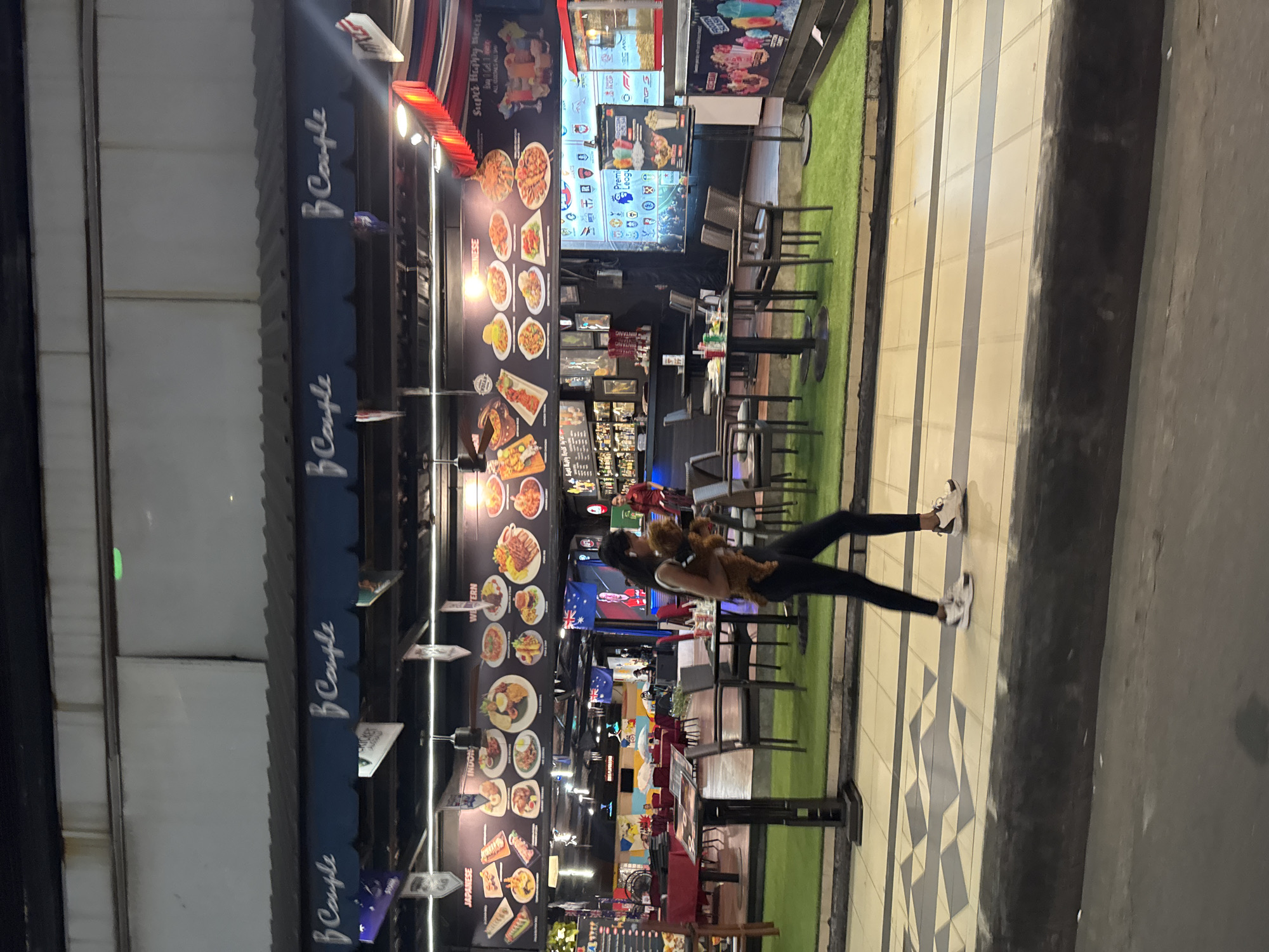 In the city of Kuta, Indonesia, a woman walks on a paved sidewalk carrying a small, brown dog. She is wearing black leggings and white athletic shoes. Behind her, an outdoor dining area is set up with tables and chairs on a green, artificial turf surface. The dining area is in front of a building with a dark awning displaying the logo "B Couple" multiple times. Above the awning, a large black banner features images of various food dishes categorized by cuisine, including Japanese, Indonesian, Western, and Chinese. The environment suggests a commercial strip with multiple food vendors or a large restaurant with diverse offerings. Flags, including an Australian flag, are visible hanging from the building. A bar area with bottles and glasses is also visible in the background. The scene appears to be at night, with artificial lighting illuminating the area.