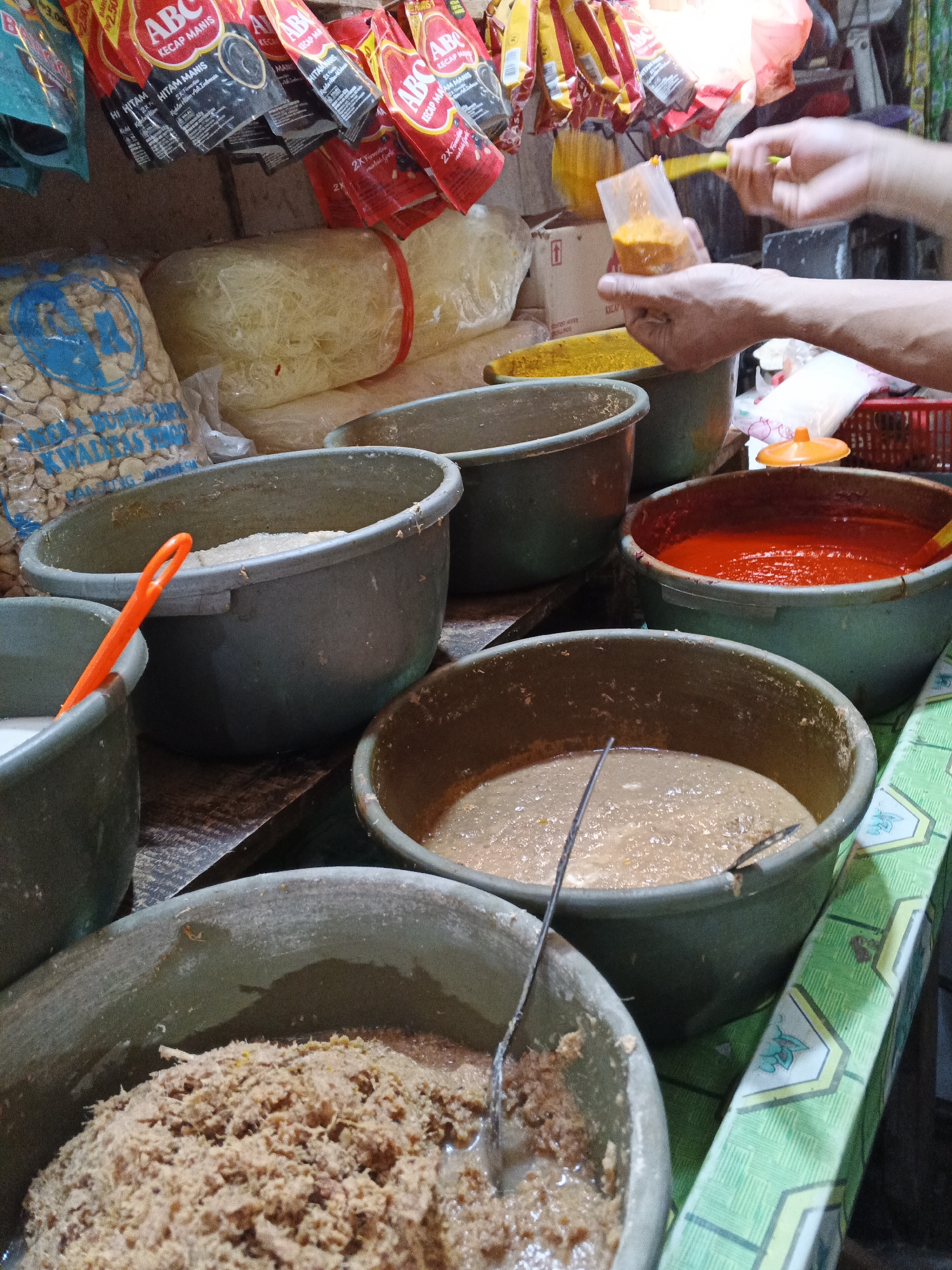 The image captures a close-up view of a food stall in Warungkiara, Indonesia, with a focus on various ingredients stored in large, dark green plastic tubs. In the foreground, a tub contains a shredded, brownish mixture, possibly meat or fish, partially submerged in liquid. To its left, another tub holds a lighter colored paste. Behind these, a tub is filled with a vibrant red sauce, and to its left, a tub contains a lighter, beige-colored paste. Further back, several more tubs are visible, some containing light-colored ingredients. Hanging above are several packets of "ABC Kecap Manis" (sweet soy sauce), indicating the stall's offerings. In the upper right, hands are visible, with one hand holding a small plastic packet filled with orange powder, and a utensil, possibly a spoon or spatula, is being used to transfer the powder. The overall environment suggests a busy marketplace or food preparation area, with wooden surfaces and a green patterned tablecloth visible.