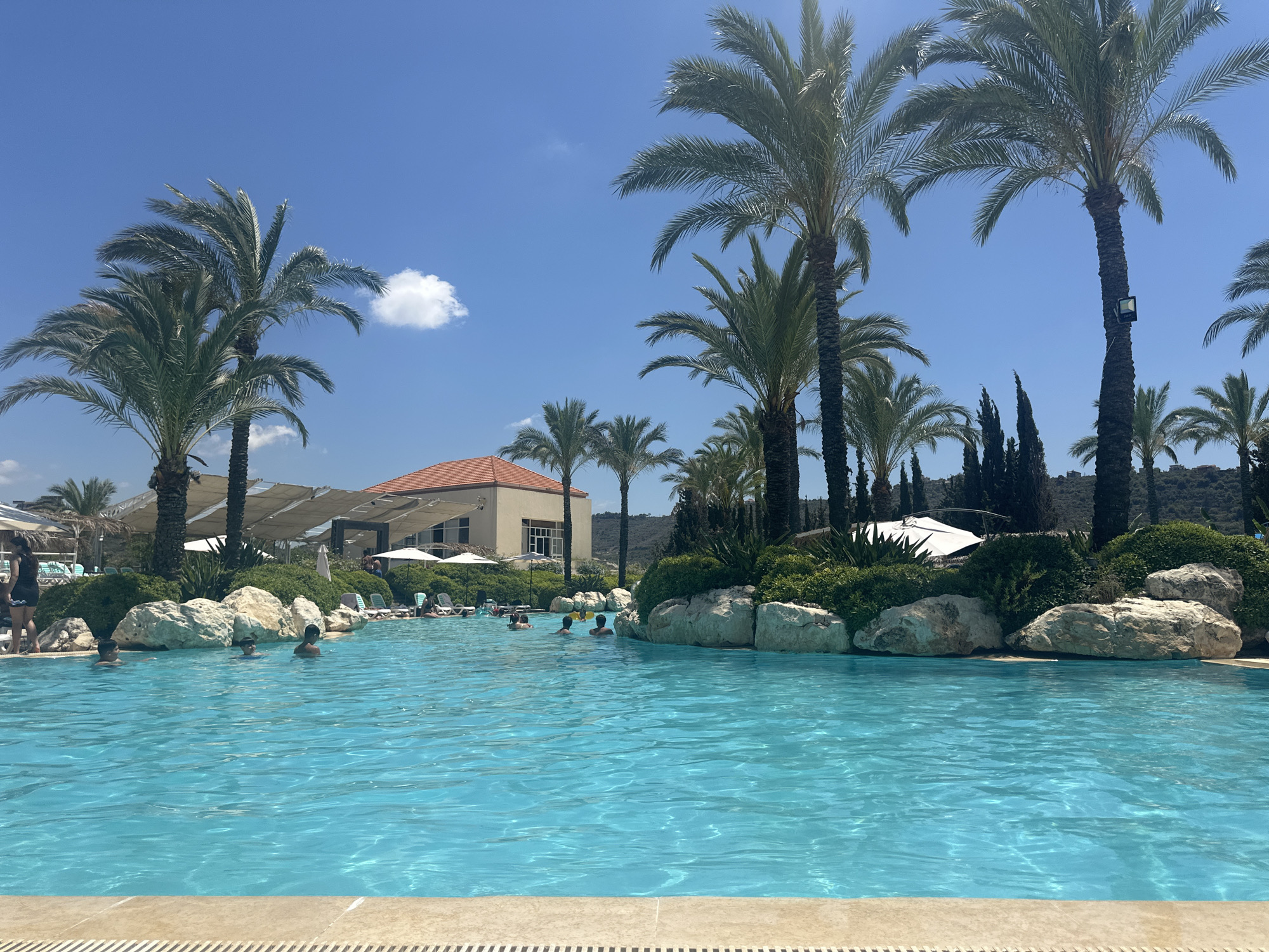 The image depicts a resort swimming pool scene in Qraiyeh, Lebanon. A large, turquoise swimming pool occupies the foreground, with its edge visible at the bottom of the frame. Several people are in the pool, some partially submerged and others swimming. The background features lush greenery, large rocks, and palm trees, characteristic of a Mediterranean resort. A building with an orange roof is visible behind the greenery, and white umbrellas and lounge chairs are scattered around the pool area, suggesting a recreational setting. The sky is a clear blue with a few scattered clouds.