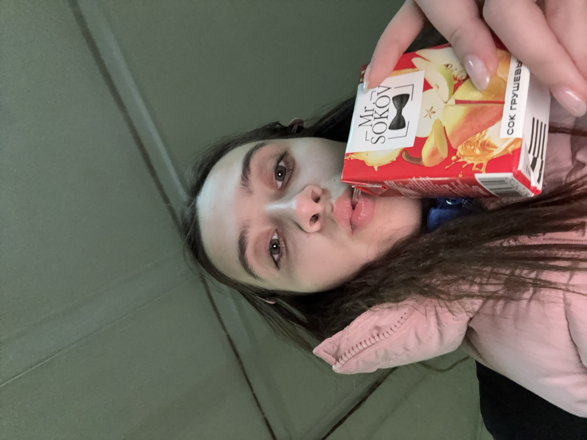 A female is holding a carton of Mr. Sokov pear juice to her lips. She appears to be indoors, with a light pink coat visible. The location is Berezniki, Russia, according to the writing on the juice carton.