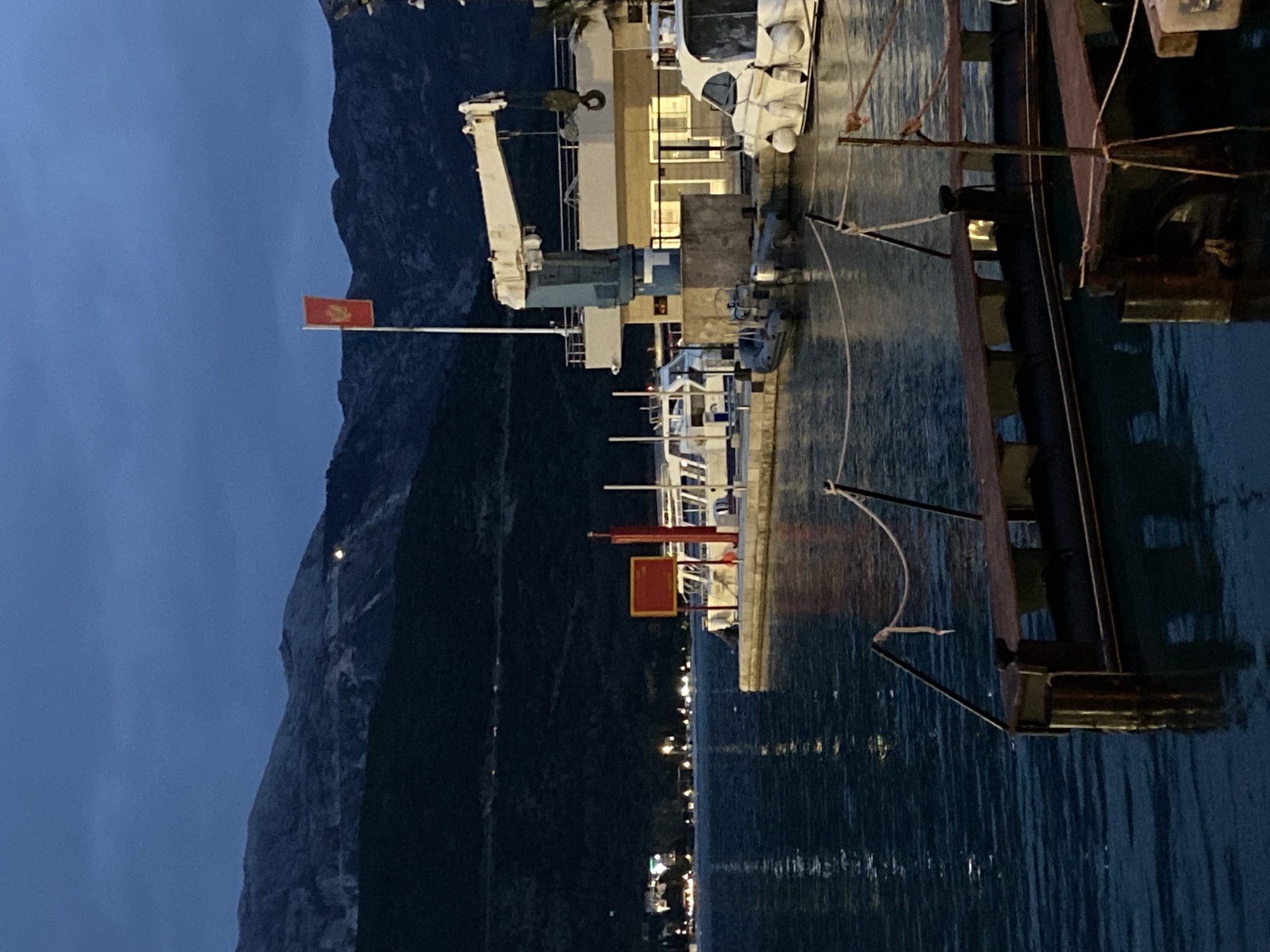 The image depicts a harbor in the city of Budva, Montenegro, at dusk or night. The left side features a dark, steep mountainside illuminated by a single light. The center shows the harbor with docked vessels. Two red flags on poles are visible. The water reflects city lights and the moored boats. A crane is seen on one of the larger vessels. A wooden dock extends into the water in the foreground, obstructing part of the view.