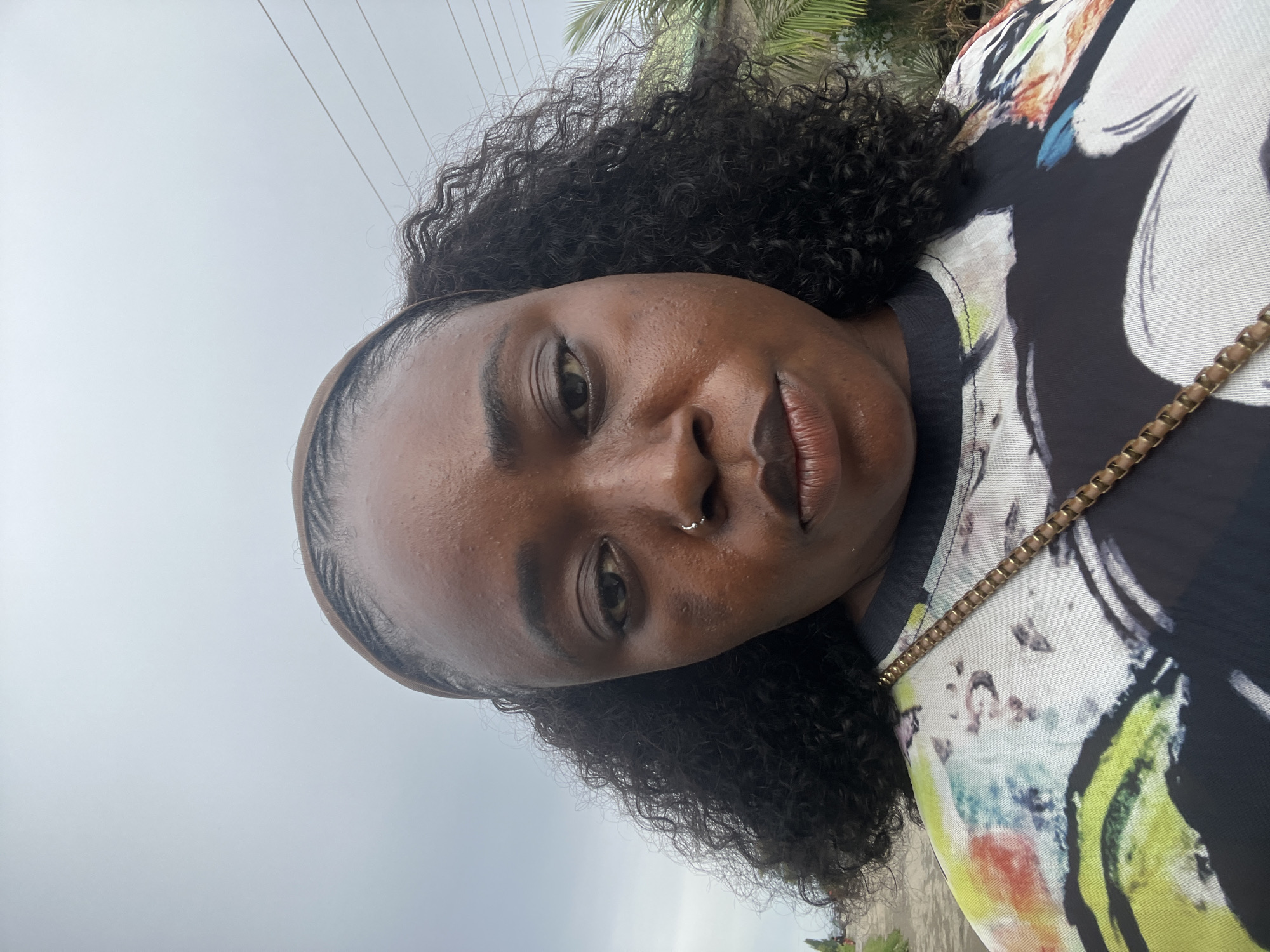 A medium shot selfie is taken outdoors in what appears to be a residential area in Abuja, Nigeria. The subject, a Black woman with dark, curly hair and a nose ring, is looking directly at the camera with a neutral expression. She is wearing a black t-shirt with a colorful, abstract pattern of green, yellow, red, and blue shapes. A gold chain strap from a bag is visible draped across her left shoulder. The background is blurred, but suggests an overcast sky with power lines, some greenery, and possibly a street or path with distant figures. The lighting is soft and diffused.