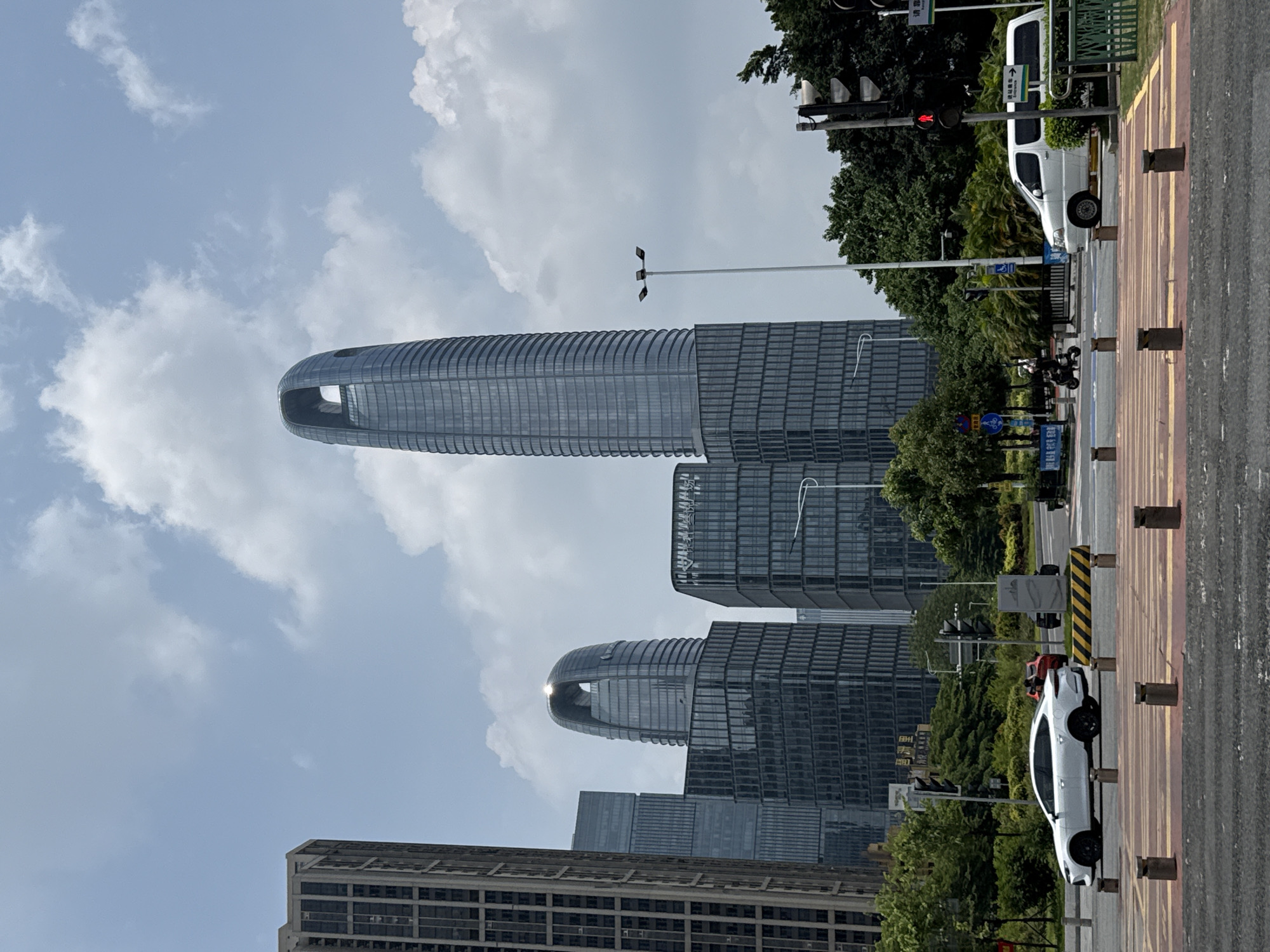 This image captures a modern urban street scene in Haizhu District, China, under a bright, partly cloudy sky. The dominant subjects are several towering, contemporary skyscrapers with reflective glass facades.
The most striking building, centrally located, is a very tall, slender skyscraper featuring a distinctive rounded, elongated "keyhole" or "bottle opener" design near its summit, tapering smoothly upwards. This unique architecture identifies it as the Guangzhou CTF Finance Centre. Below it, a similar but shorter high-rise is visible, alongside other rectangular glass buildings in the background, contributing to a dense metropolitan feel.
At street level, a multi-lane road system is visible, composed of asphalt and sections of red and yellow paving. A series of brown bollards lines the paved sidewalk area. Street furniture includes tall streetlights, a traffic light pole displaying a red signal, and various signs with Chinese characters. A white commercial van is parked near a curb on the right side of the road, and a sleek white car with dark wheels is parked closer to the foreground. A motorcycle or scooter is also visible further down the street. Abundant green trees and bushes line the street and punctuate the spaces between buildings, adding natural elements to the concrete and glass environment.
The time of day appears to be late morning or early afternoon, characterized by bright sunlight, casting reflections off the glass buildings and creating a clear blue sky with scattered white clouds. No people are prominently visible, and the scene depicts a static, functional urban landscape. Visible text on signs is in Chinese, aligning with the specified location context.