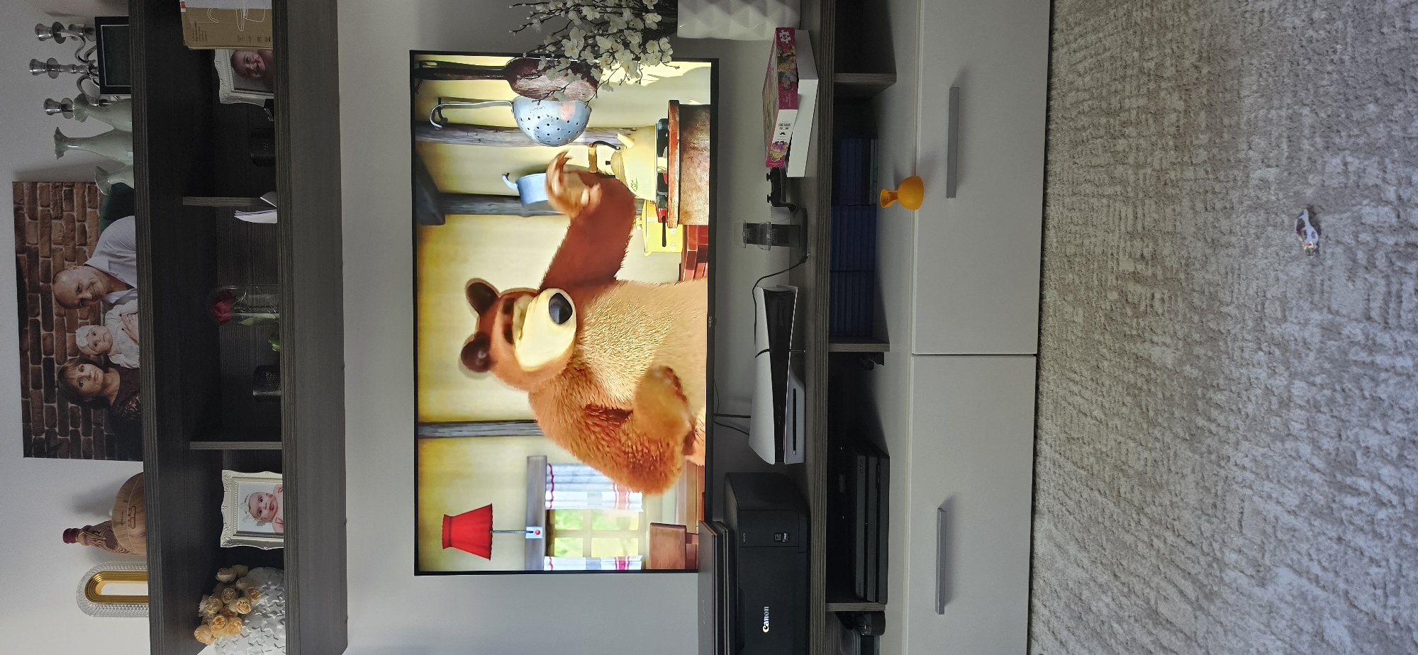 A television screen displays a scene from the animated series "Masha and the Bear," featuring a large brown bear in a kitchen setting. The bear is holding a teacup in its right paw and is looking towards the viewer. To the left of the bear, a red lampshade hangs from a wall. The screen is placed on a dark gray shelf, above a white and gray media console. On the media console, a black Canon printer is visible on the left, and a white PlayStation 5 console is positioned to the right of the printer. A stack of blue game cases is stored below the PlayStation 5. A small orange toy duck rests on the edge of the media console. The room's walls are white, and a carpet with a textured pattern covers the floor. Above the television, on a dark gray shelf, there are several decorative items including framed photographs of a family, vases, and candlesticks.