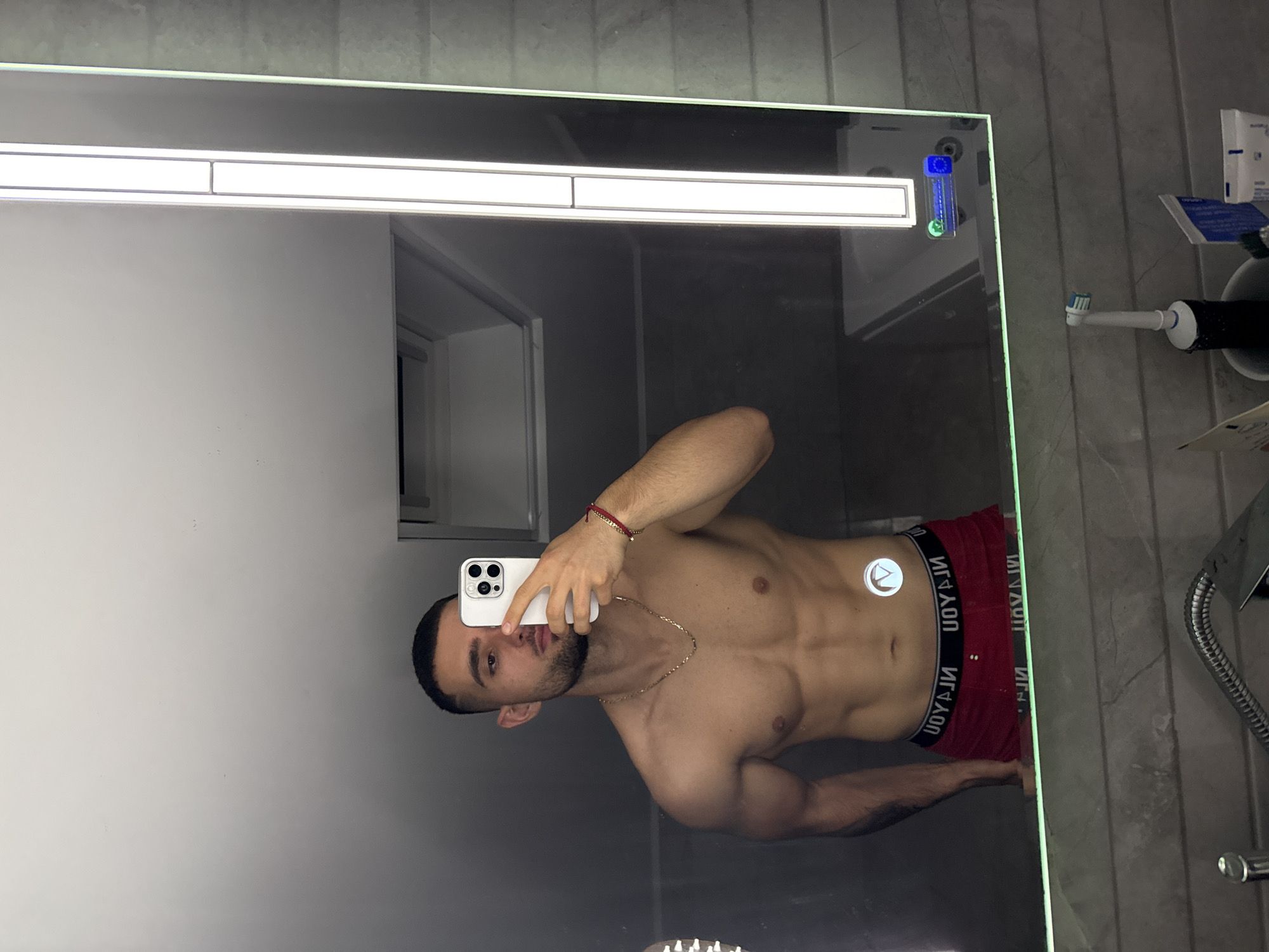 A shirtless man is taking a selfie in a mirror. He is wearing red underwear with a black waistband. He has a beard and is wearing a gold chain. A white smartphone is visible in his hand. A light fixture is above the mirror, and a toothbrush and other toiletries are on the counter to the right. The location is a bathroom. The image doesn't have enough information to determine if it is in the city of Gabrovo, Bulgaria.