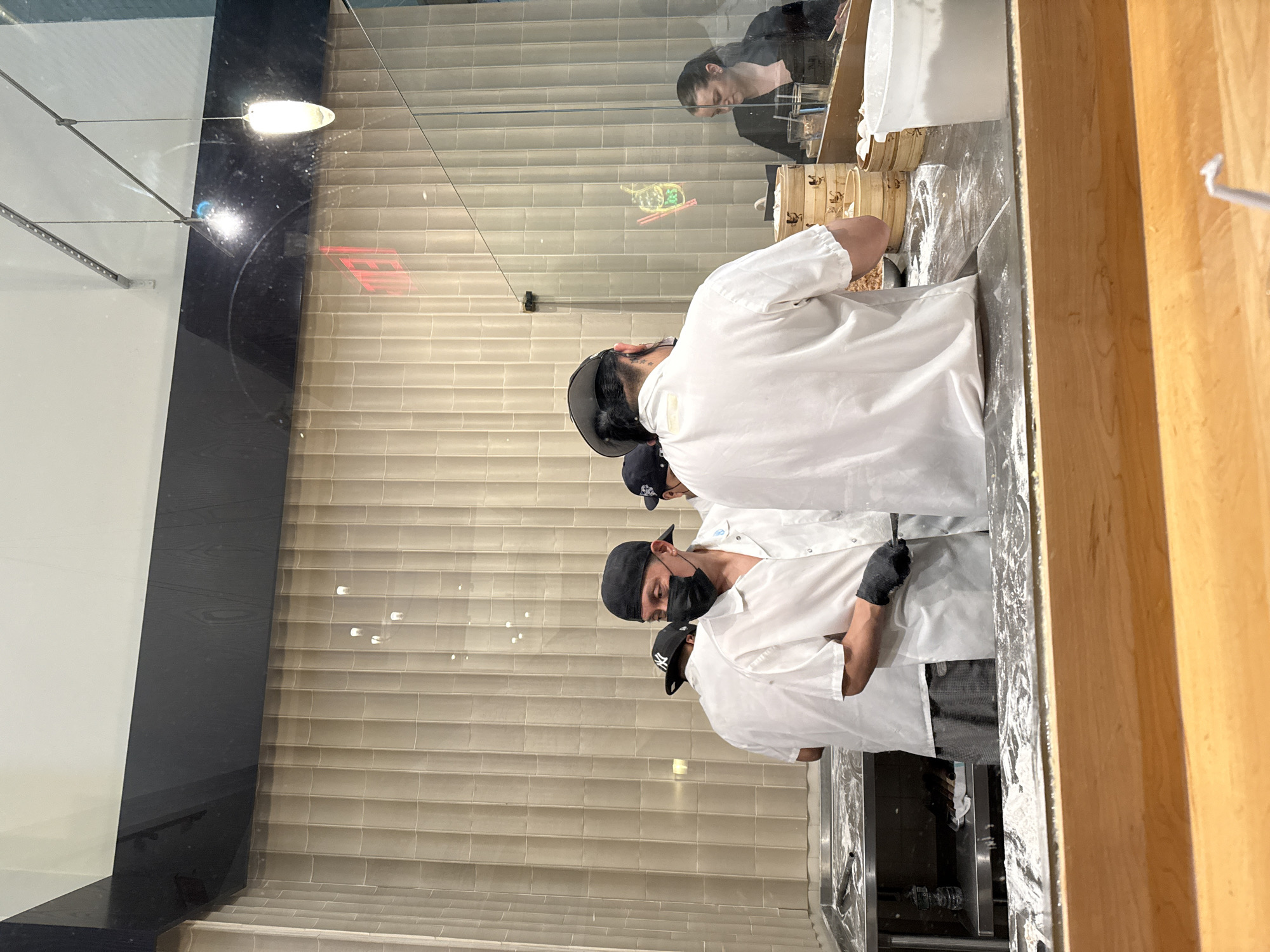 The image depicts a culinary scene, possibly in New York City, United States of America, as suggested by the architectural style of the building and the commercial nature of the setting. Three individuals are present, all wearing white shirts, black gloves, and black baseball caps. The person nearest the camera is wearing a face mask. They are positioned behind a counter or workstation, which is covered in flour, and appear to be engaged in food preparation.
The background features a tiled wall and a glass partition, through which a reflection of another individual can be seen. This person appears to be seated and possibly observing the food preparation process. The overall setting suggests a restaurant or a food preparation area open to public view.