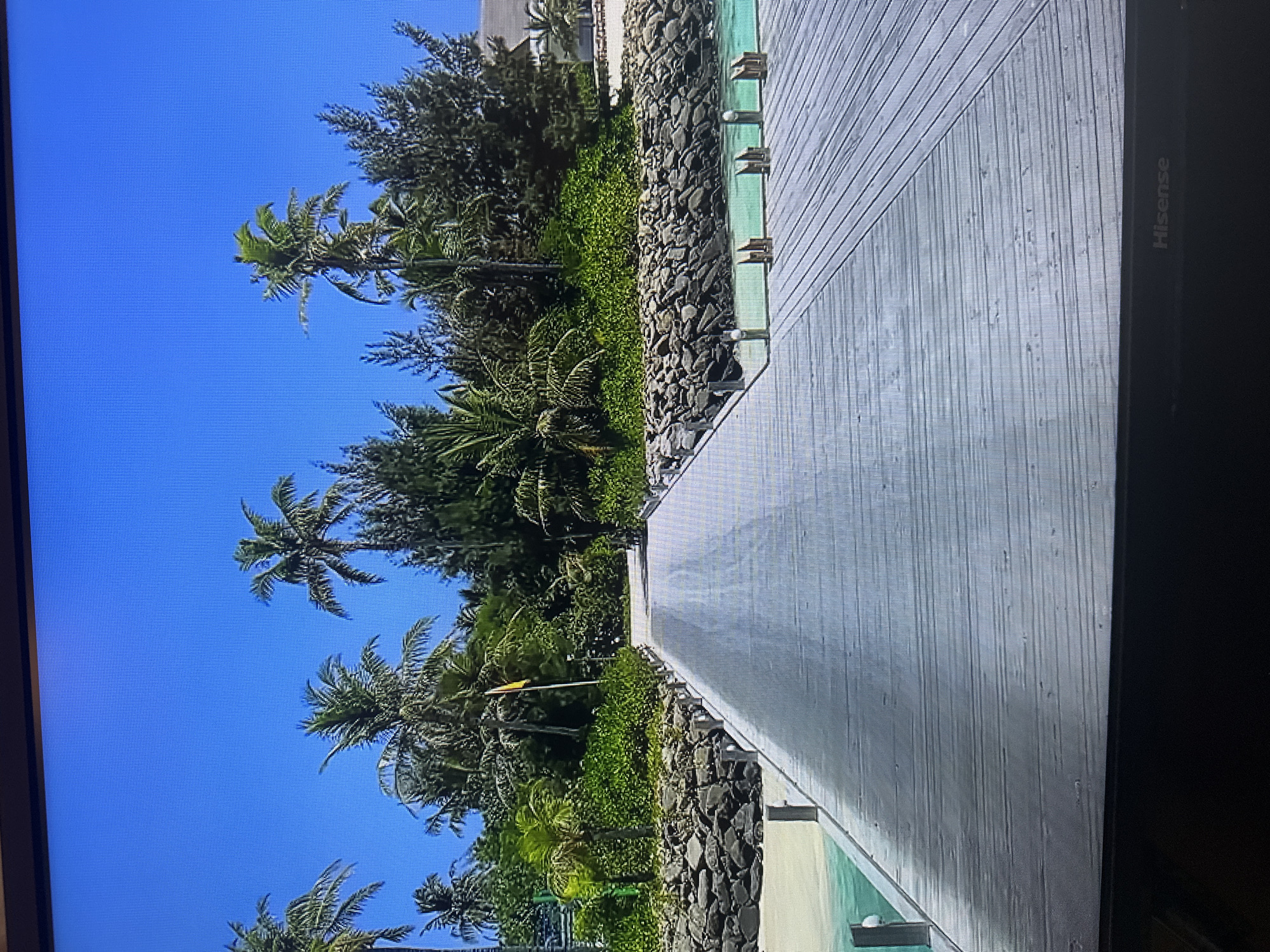 The image depicts a wooden pier extending into clear turquoise water under a bright blue sky. Palm trees and lush green vegetation are visible on the shore, with a rocky embankment lining the water's edge. The pier has a ramped section leading upwards and appears to be made of weathered wooden planks. In the distance, part of a building with a light-colored roof is visible among the trees. The scene evokes a tropical island or resort. The location is not in Kyiv, Ukraine.