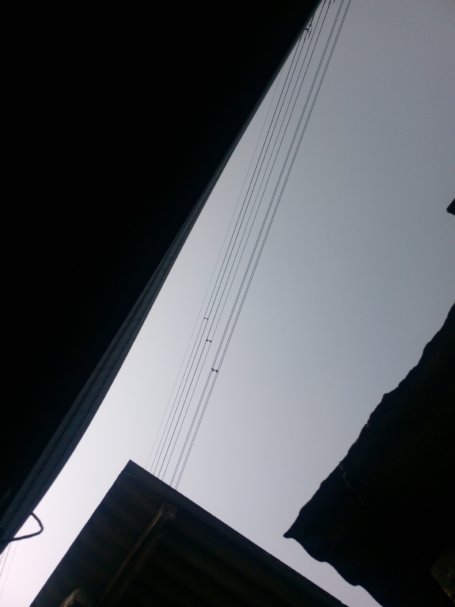 The image captures a low-angle view of the sky in Dukuh, Indonesia, framed by dark, silhouetted structures. Several power lines are visible, stretching diagonally across the pale sky. The foreground features the edge of a dark, angular structure on the left, and another jagged, dark silhouette on the right, possibly the edge of a roof or building. In the lower center, a portion of a dark, wooden roof structure is visible. The overall impression is of an urban or semi-urban environment, looking upwards from a confined space.