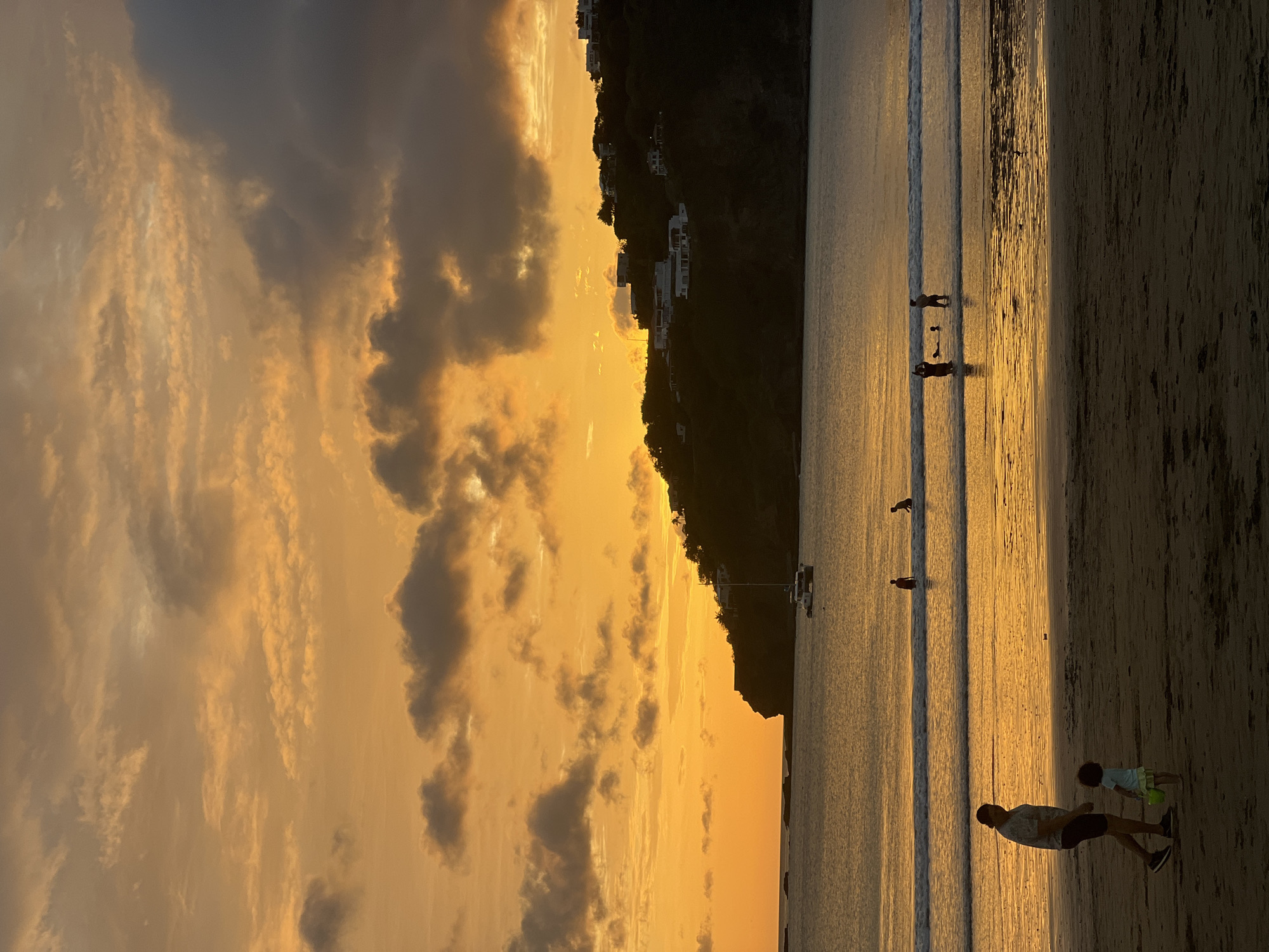 This image depicts a sunset over the Pacific Ocean at San Juan del Sur, Nicaragua. The sky is filled with vibrant orange and yellow hues, with scattered dark clouds. In the foreground, a sandy beach is visible with a man walking and a small child playing with a yellow bucket. The man is wearing a patterned shirt and dark shorts, while the child is wearing a light-colored dress. Further out, in the shallow water, several people are wading and playing in the gentle waves. A sailboat is anchored in the bay, and on the dark, tree-covered hillside in the background, several white houses are perched, silhouetted against the colorful sky. The calm water reflects the warm colors of the sunset.