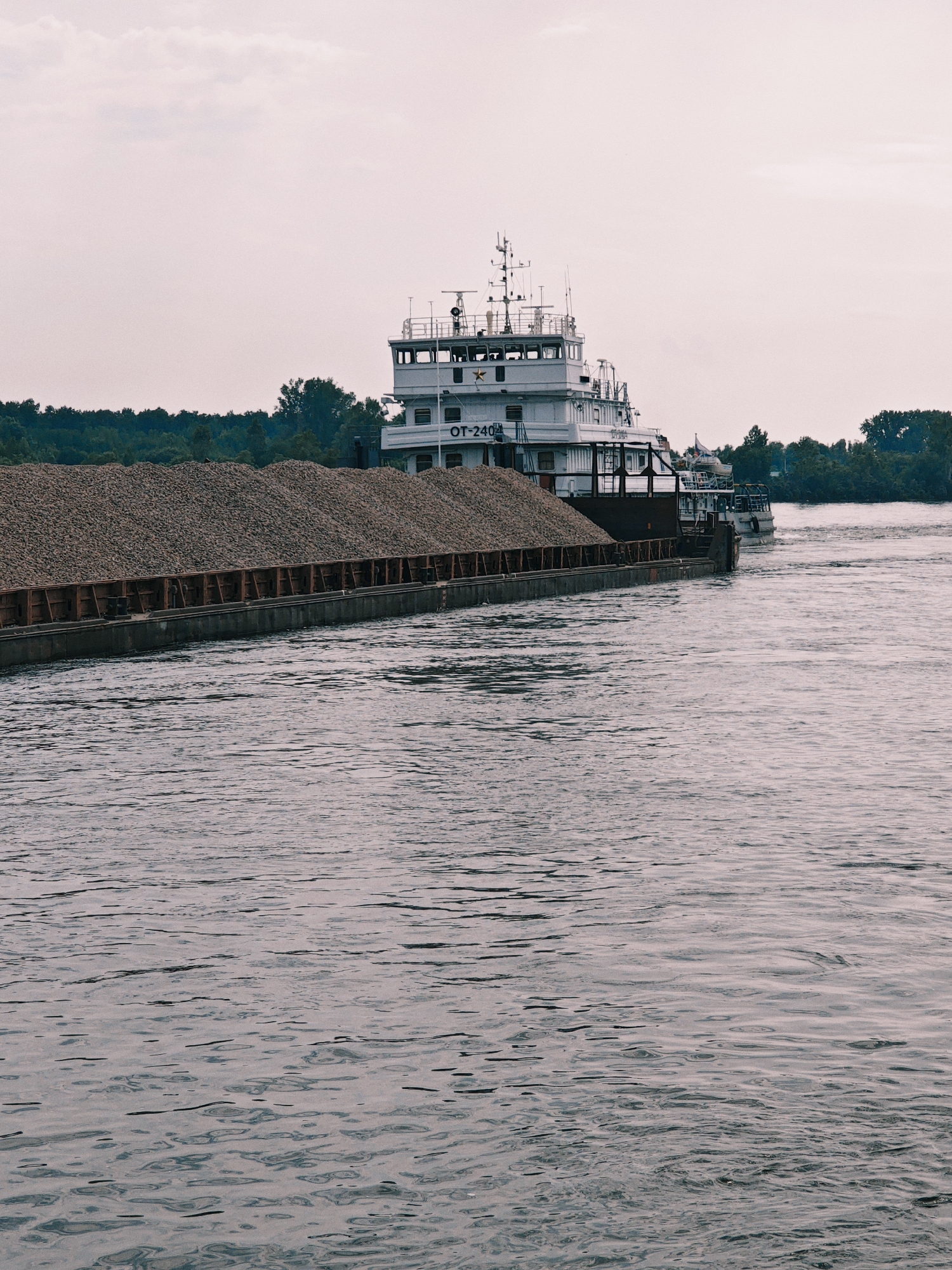 An overcast day on the Yenisei River near Sosnovoborsk, Russia. A large barge, laden with a substantial cargo of what appears to be gravel or crushed stone, is moving through the water. The barge has a prominent superstructure with the designation "OT-240" visible. To the right, a smaller tugboat is pushing the barge. The riverbanks are lined with dense green trees. The water is choppy, with visible ripples and wakes. The sky is a pale, muted gray.