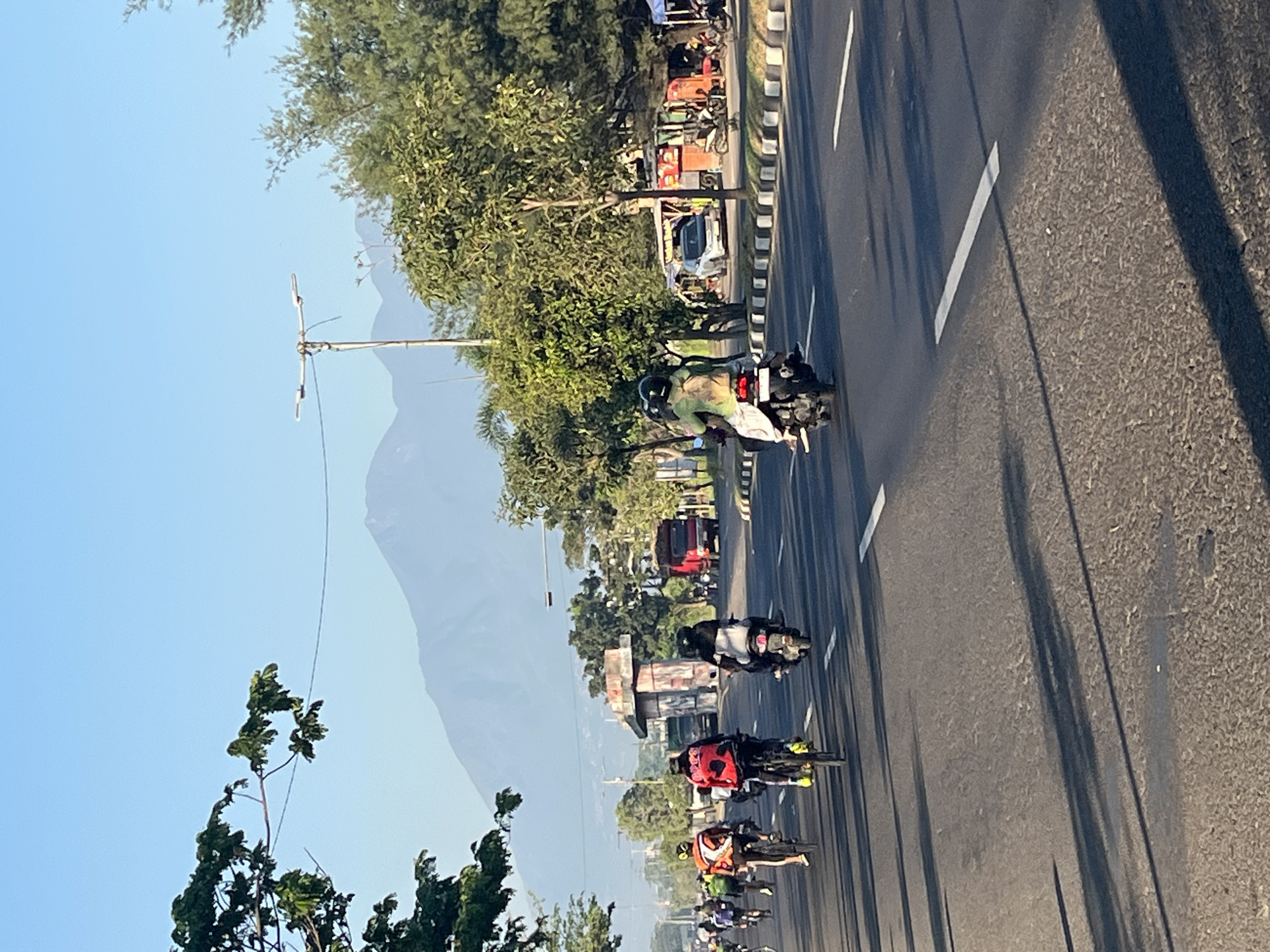 In Siring, Indonesia, a bright, clear morning showcases a road filled with cyclists and a few motorcycles. In the foreground, a group of cyclists are riding on the right side of the road, their colorful jerseys visible. To the right, a motorcycle carries two individuals, also heading in the same direction. The road is asphalt and marked with white lines. On the left side of the road, trees line the shoulder, with some branches extending over the road. In the background, a red truck and other vehicles are visible, along with a large concrete structure that appears to be a construction or underpass support. Beyond that, a hazy mountain range dominates the horizon under a clear blue sky. Shops and stalls line the right side of the road in the mid-ground, with more parked motorcycles. The sun casts long shadows across the road, indicating early morning.
