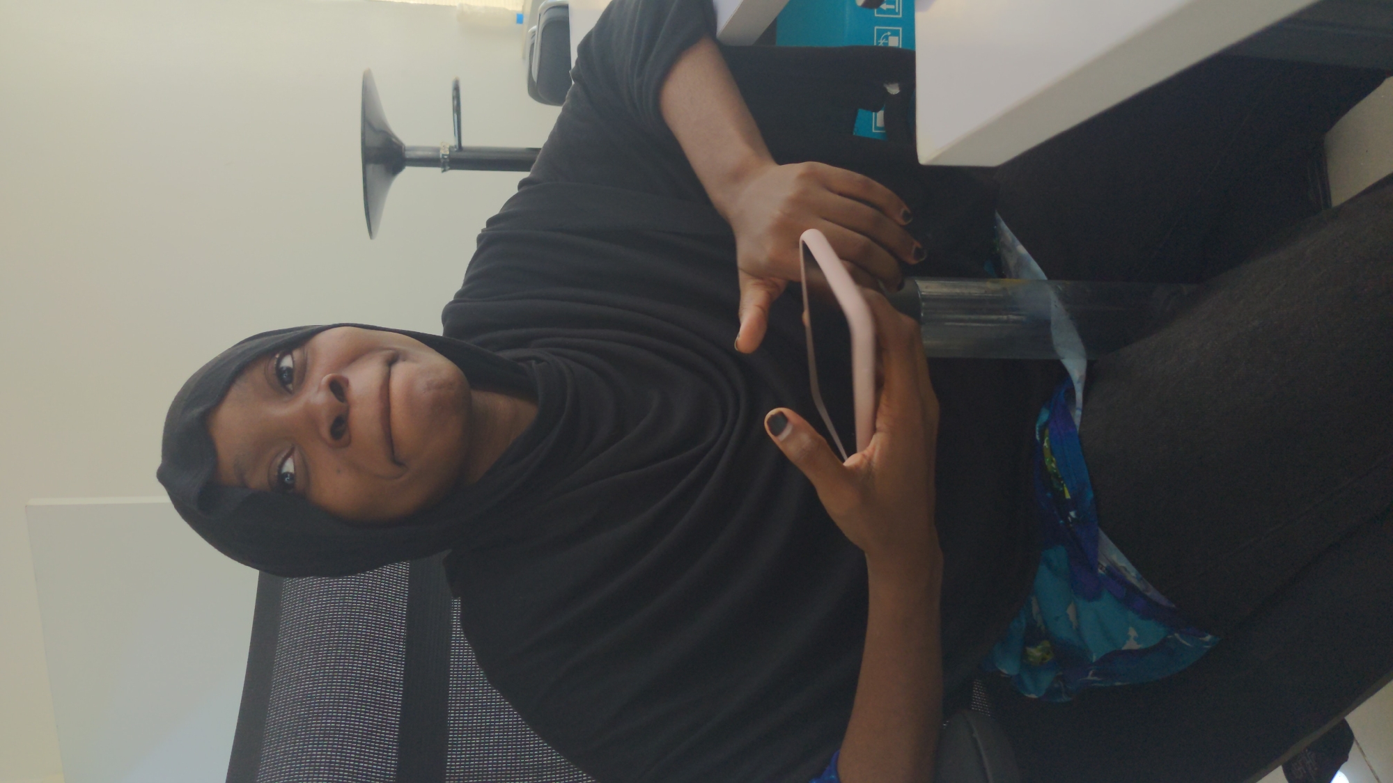 An indoor, eye-level shot captures a person, likely female, with dark skin, seated in an office-like setting. She is looking directly at the camera with a playful or slightly mischievous expression, featuring slightly puffed cheeks and a subtle smirk. Her head is covered by a black hijab or headscarf, and she wears a long-sleeved black top. Her fingernails are painted with dark polish.
She holds a light-colored (possibly pink or beige) smartphone in both hands. She is positioned in a black office chair with a mesh back. Her lower body is clad in dark trousers, and a blue and white patterned scarf or fabric is draped over her lap. To her right, a white desk or table surface is visible. The background consists of a plain, light-colored wall. A black, trumpet-shaped stand, possibly part of a lamp or microphone, extends from the background wall. A blue and white object, which appears to be a box with faint packaging symbols (like arrows), is partially visible behind the desk. The scene depicts a casual moment in what appears to be a contemporary workspace. No discernible text is visible.
