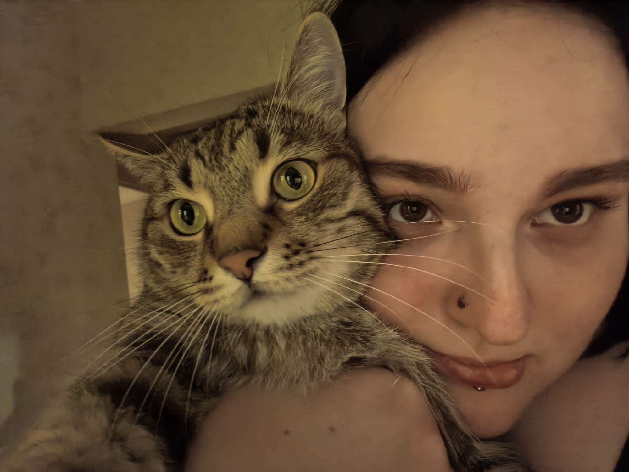 A close-up selfie shows a person holding a tabby cat. The person's face is partially visible on the right side of the frame, with their eyes looking at the camera. The cat is held close to the person's face, with its head and upper body in the left portion of the image. The cat has green eyes and striped fur. The background is a plain, softly lit wall.