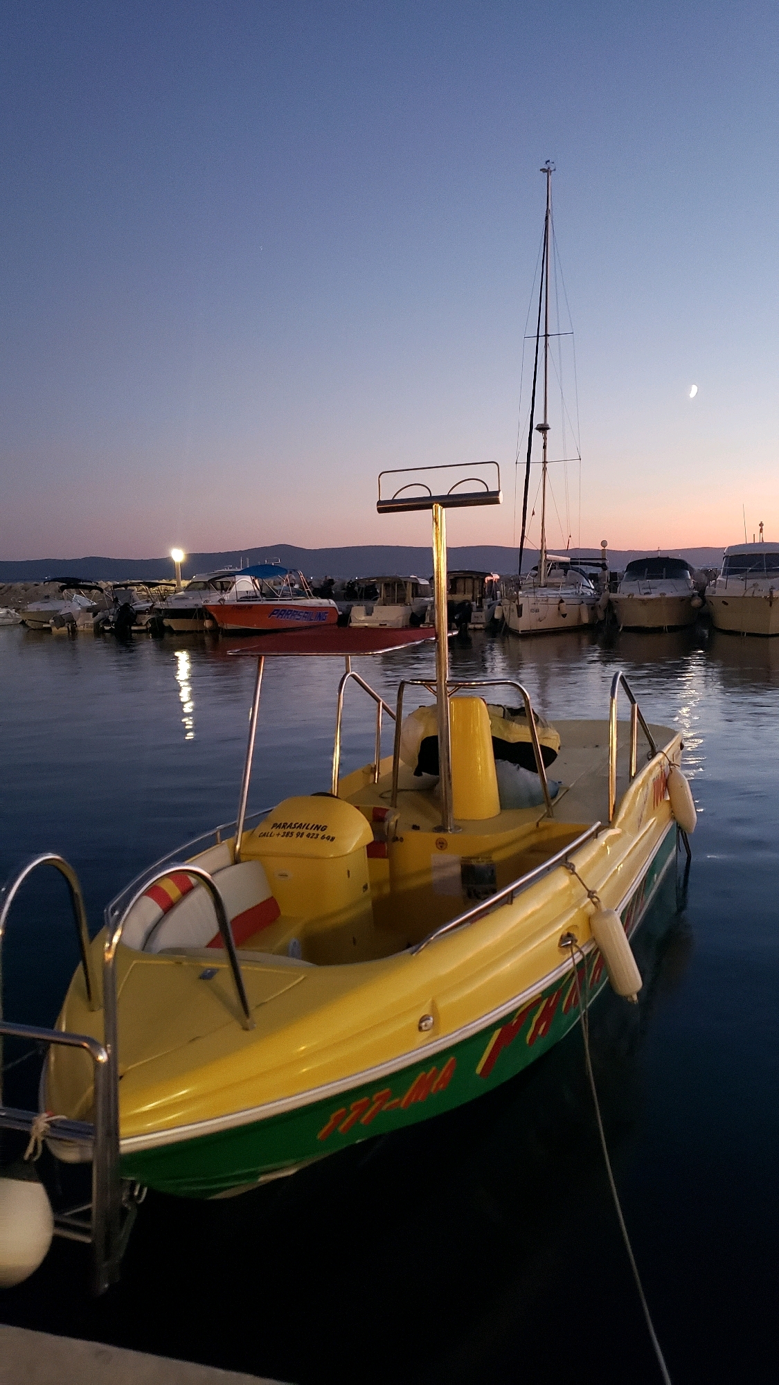 The image shows a marina in Općina Tučepi, Croatia at dusk. The sky is a gradient of deep blue to a light pinkish-orange near the horizon, with a crescent moon visible. Several boats are docked in the water. In the foreground, a bright yellow boat with green trim is prominently featured. It has a life preserver on the left side and a metal frame with two curved bars at the top. Text that appears to be "PARASAILING CALL +385 98 423 648" is visible on a yellow seat in the boat. In the middle ground, other boats are docked, including a red boat with "PARASAILING" written on its side. In the background, a sailboat with a tall mast is visible, along with more docked boats and a distant coastline with mountains. The water is calm and reflects the lights from the marina. The overall scene is peaceful and suggests the end of a day's activities.