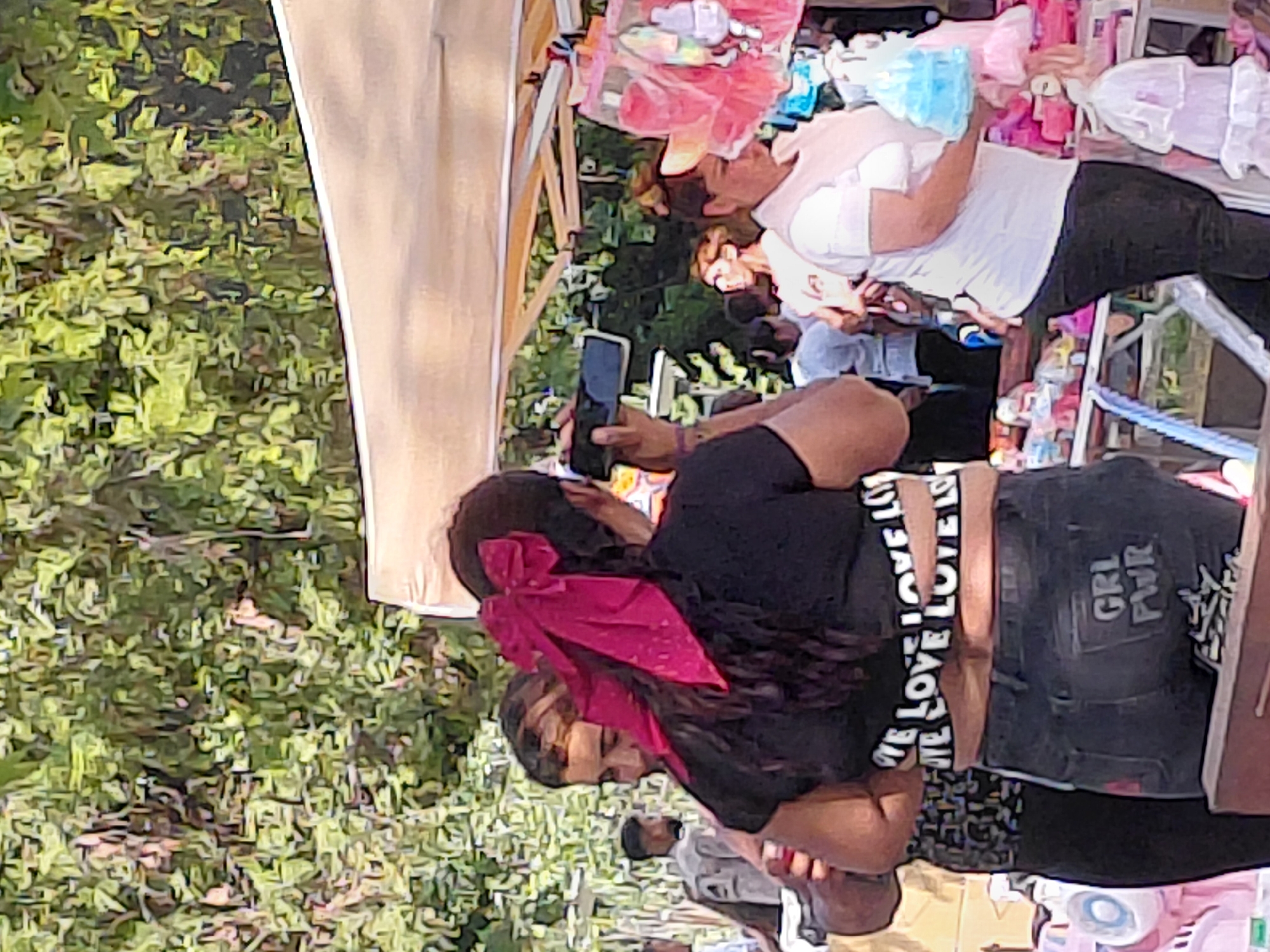 An outdoor scene, likely a market or festival, captured in the city of Kostievo, Bulgaria. In the central foreground, a woman stands with her back partially to the viewer, exhibiting long, dark hair tied up and adorned with a prominent magenta bow. She wears a black crop top featuring "LOVE LOVE LOVE LOVE LOVE" repeated in white text across the midriff band, and dark jeans with "GRL PWR" (Girl Power) printed in white on the right rear pocket. This woman is actively holding a black smartphone horizontally with both hands, appearing to photograph or record something in front of her. Her left arm is around another individual, partially visible on the left, who has dark hair and is wearing a patterned dark top.
To the mid-right, another woman is observed in profile, facing left, with dark hair and a light-colored cap or visor. She is dressed in a white short-sleeved top and dark trousers, and appears to be examining or holding light-blue ruffled garments, possibly for dolls or children, which are part of a display. Various other pink and white ruffled items are suspended from a market stall structure, visible behind her.
In the mid-background, to the left of the central figures, a man in a grey long-sleeved shirt with a logo (partially obscured) on his left arm is seen with his back to the camera, standing near other indistinct figures. The background is dominated by dense, light-green foliage, indicative of trees or bushes, suggesting a natural outdoor setting. A light-colored market canopy extends across the upper right portion of the image. The immediate foreground shows a dark, flat surface, potentially a counter or table. The overall scene suggests a busy, daytime event with multiple people and merchandise displays.
