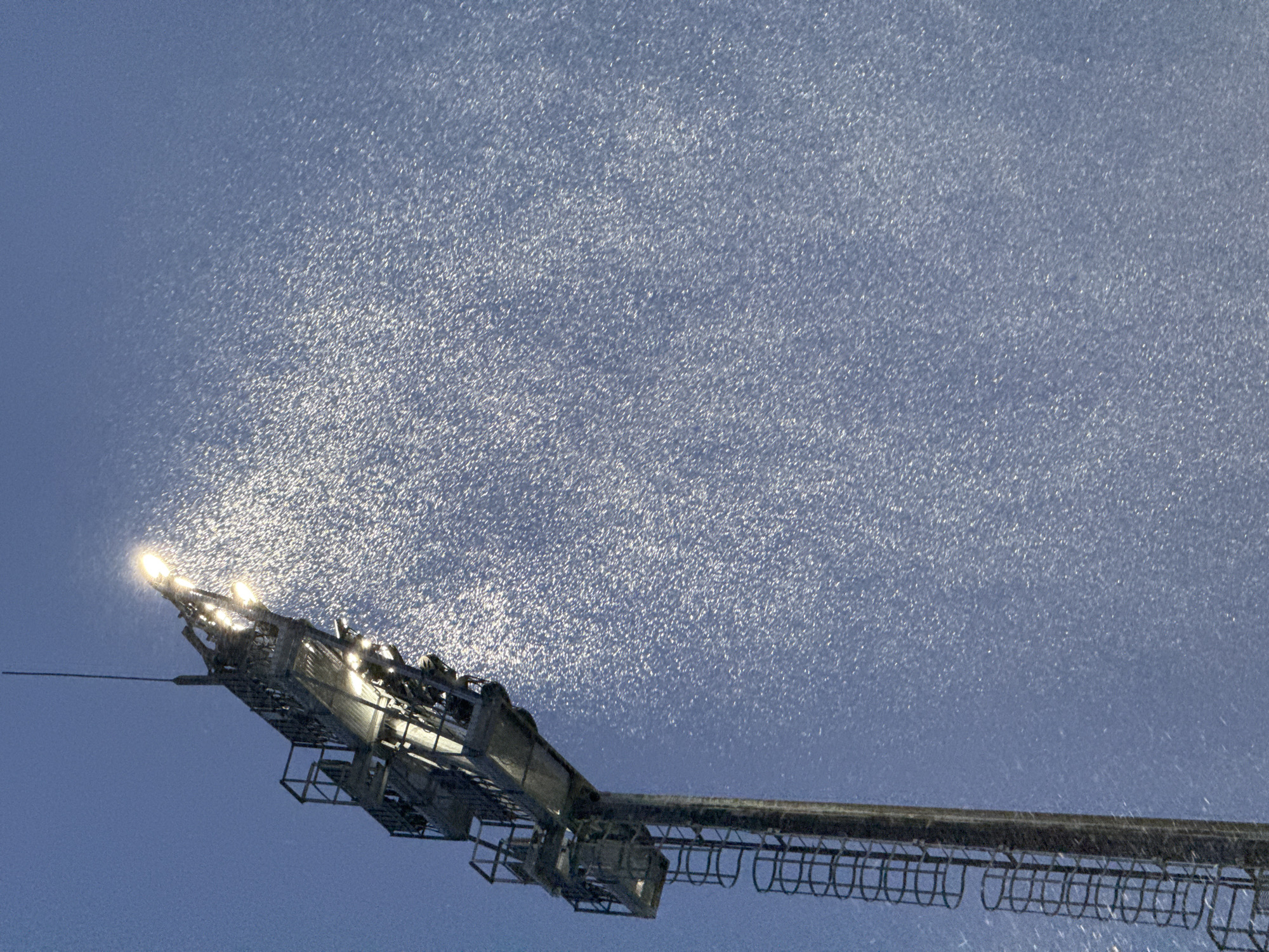 The image displays a stadium light tower against a deep blue, dark sky, likely at night in Moscow, Russia. The prominent subject is the end section of a large, metallic structure, fitted with numerous bright floodlights that illuminate the surrounding atmosphere.
Thousands of small, shimmering white particles are visible in the air, caught and highlighted by the powerful light beams. These particles appear to be falling precipitation, most likely snow or sleet, creating a dense, sparkling curtain around and above the light source. The intensity of the lights causes these particles to glow brightly, giving the scene a granular, almost textured appearance.
The light tower itself is a complex assembly of metal beams and cages, extending from the lower right corner and angling upwards, with its brightest part at the top left where the main lights are situated. The sky transitions from a lighter, illuminated blue near the lights to a darker, more muted blue further away.
The overall impression is one of a cold, wintry night, possibly during a sporting event or outdoor activity where such floodlighting is essential. The weather conditions are clearly visible due to the interaction of light with the atmosphere. No discernible text is present in the image.