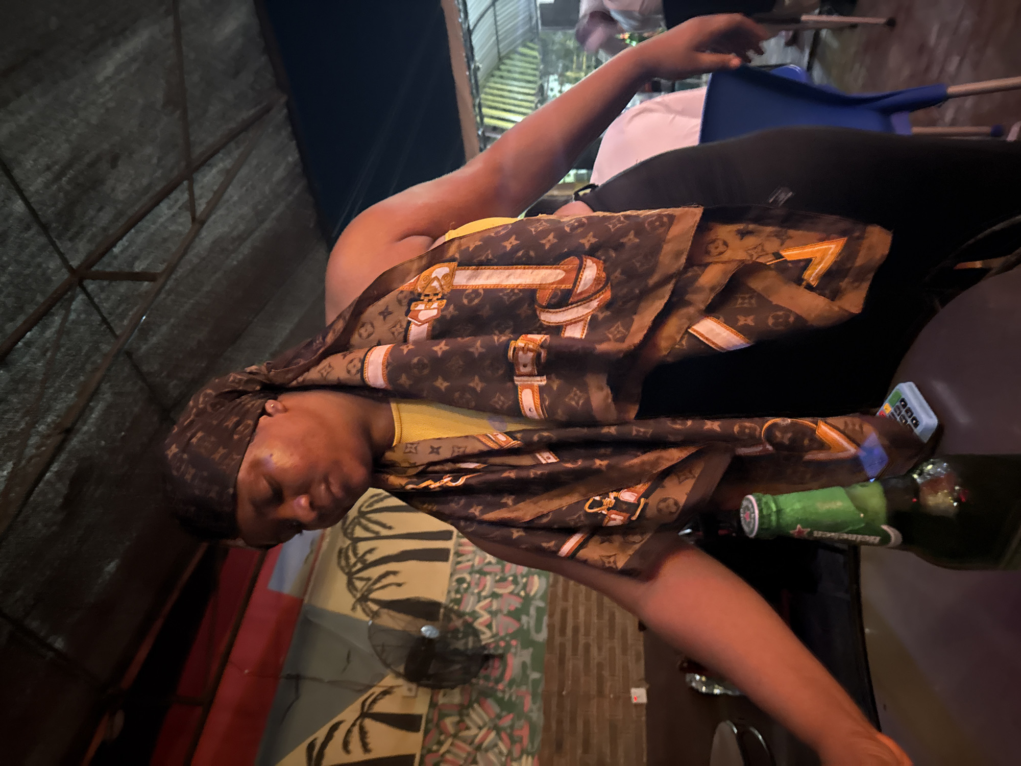 This image depicts an indoor or semi-outdoor social gathering space, likely a bar or restaurant, located in the city of Ado, Nigeria, at night. The primary subject is a woman of African descent, shown from the waist up, leaning slightly forward and to the left. She wears a yellow sleeveless top, black trousers or leggings, and a brown scarf with a repeating pattern resembling Louis Vuitton monograms and belt/buckle designs. The scarf covers her head and is draped over her left shoulder. Her head is tilted downwards, and her eyes appear partially closed. Her left arm extends towards the left foreground, out of frame, while her right hand is visible near her hip.
In the immediate foreground, on a dark table surface, stands an uncapped green Heineken beer bottle, next to a small, light-colored electronic device. The background features a painted wall with black silhouettes of palm trees on a light yellow section, above a colorful geometric pattern in shades of green, red, and blue, which rests on exposed brickwork. A black wall-mounted fan is visible on the left wall. The overhead structure consists of a dark, corrugated metal roof or awning supported by metal beams. The scene is dimly illuminated by artificial light, and blurry figures of other individuals and blue plastic chairs are faintly visible in the background.
