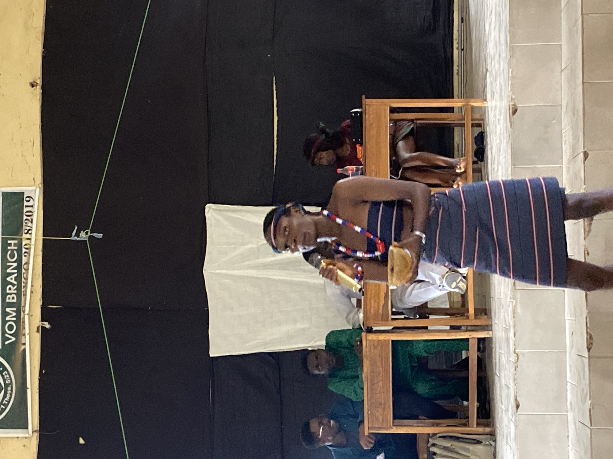 The image depicts an indoor scene with several people, likely in Nigeria given the location context. In the foreground, a young person wearing a striped skirt and a beaded necklace is holding a microphone and what appears to be a small instrument or prop. They are looking slightly to the side. Behind this individual, a white cloth is draped, and further back, another person is visible sitting, partially obscured by a wooden structure. To the left of the foreground subject, a sign with text is partially visible. The text reads "VOM BRANCH" and some numbers. The setting appears to be a stage or performance area, with a dark backdrop. The lighting suggests an indoor event. The mood seems to be attentive, possibly during a performance or presentation. The overall scene is dimly lit, but the focus is on the individuals and the activity taking place.