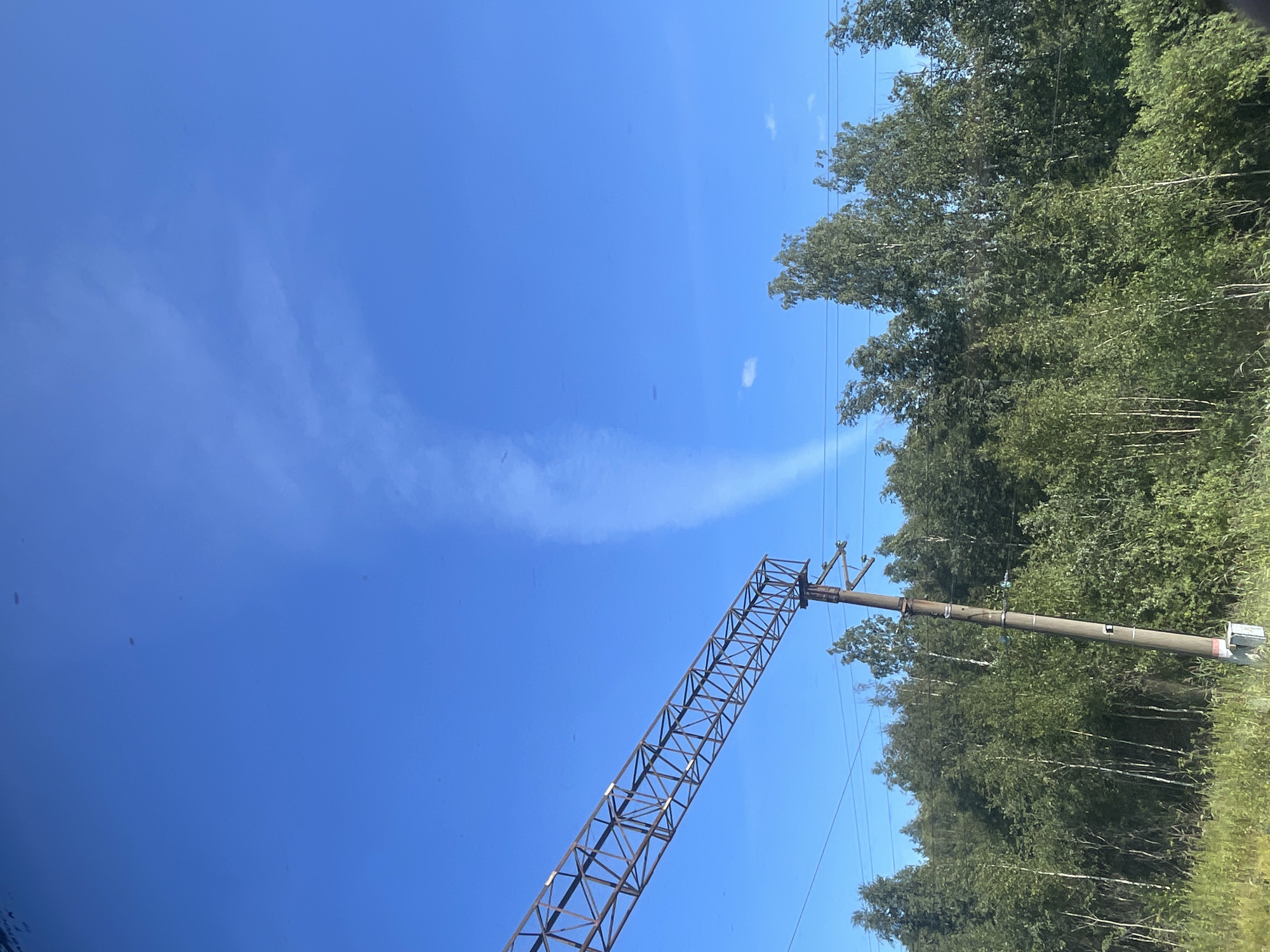A clear, bright blue sky dominates the upper portion of the image, with a thin, wispy cloud formation resembling a funnel cloud or contrail extending vertically from the upper right towards the center. In the foreground, a dense line of green deciduous trees and tall grasses creates a natural barrier across the lower part of the frame. To the left, a tall, weathered concrete utility pole stands prominently, supporting electrical wires and a large, rusty metal structure that extends diagonally across the sky, likely part of a railway overhead catenary system. The overall scene suggests an outdoor, rural or semi-rural environment on a sunny day.