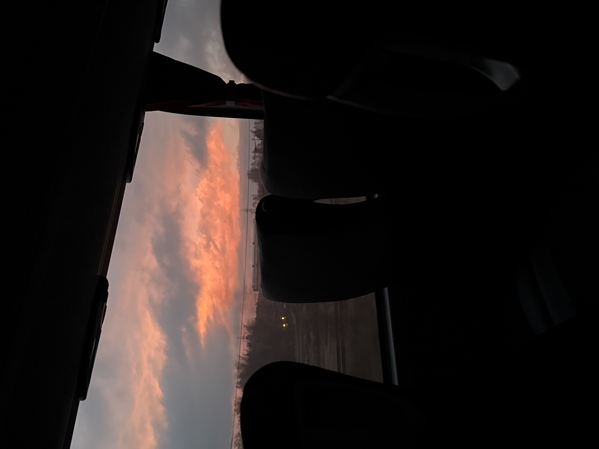 View from a bus window at dusk or dawn. The interior of the bus is dark with seats in silhouette. The window shows a landscape with a reddish-pink sky, trees, and a road. Two lights are visible in the distance, possibly from a vehicle. The image appears to be taken in Iksha, Russia.