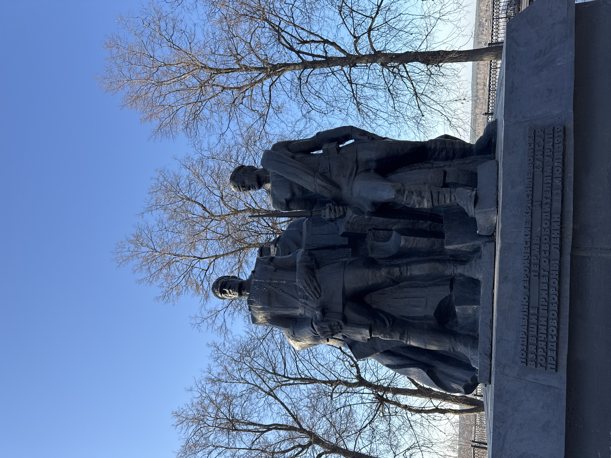The image shows a monument in Perm, Russia. The monument depicts two soldiers standing on a dark stone plinth with bare trees surrounding it. The inscription on the plinth reads: "I congratulate the heroic Red Army, Perm taking Kungur. A hot greeting to the liberators of the Urals Chairman of the Council of Defense Lenin 1 July 1919." A bright blue sky is visible in the background.