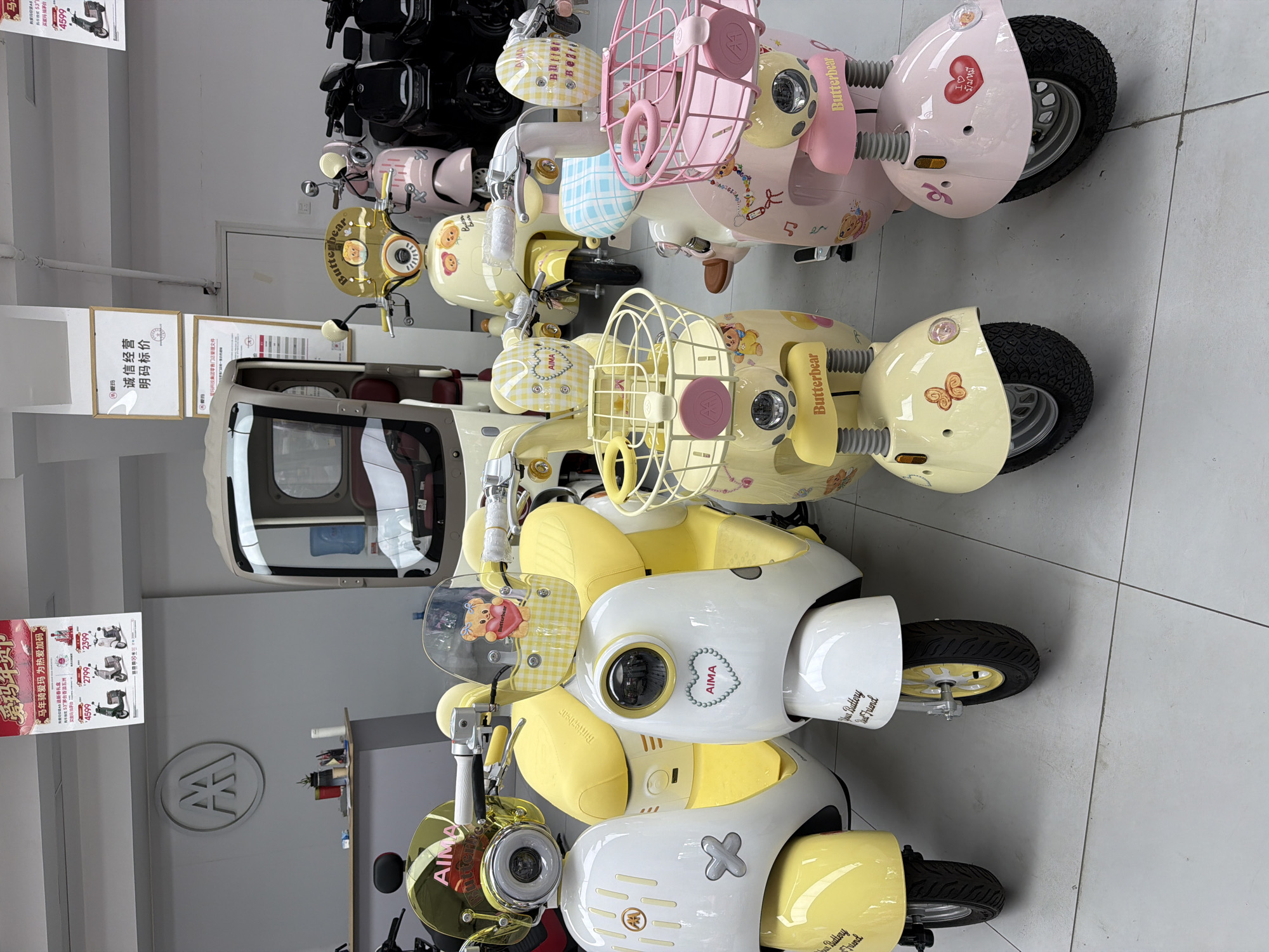 The photograph shows a retail space in Chongqing High-tech Zone, China, displaying a variety of "AIMA" brand electric scooters, designed with cartoon character themes like "Butterbear." The scooters vary in color, including white, yellow, and pink, and feature design elements such as front baskets, custom seats, and decorative motifs. The environment includes display signage with pricing, a retail counter, and other scooters in the background, indicating a showroom or dealership setting. The floor is tiled and the lighting is bright, suggesting an organized and modern commercial space.