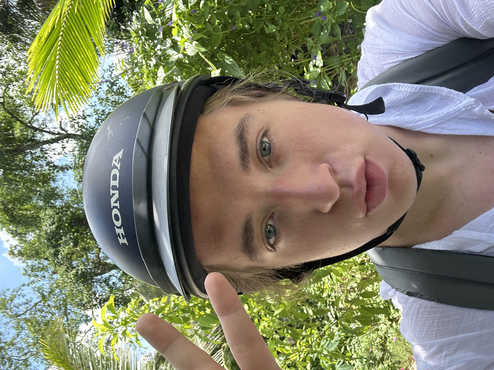 A young person with light-colored hair and blue eyes is wearing a black Honda helmet and a white crinkled shirt. They are making a peace sign with their left hand and looking at the camera. The background is filled with lush green foliage and some small purple flowers under a bright blue sky. The image appears to have been taken outdoors, possibly in a tropical environment, and the context suggests a location in An Hữu, Vietnam, given the presence of dense vegetation and the type of helmet commonly used for motorbikes in the region.