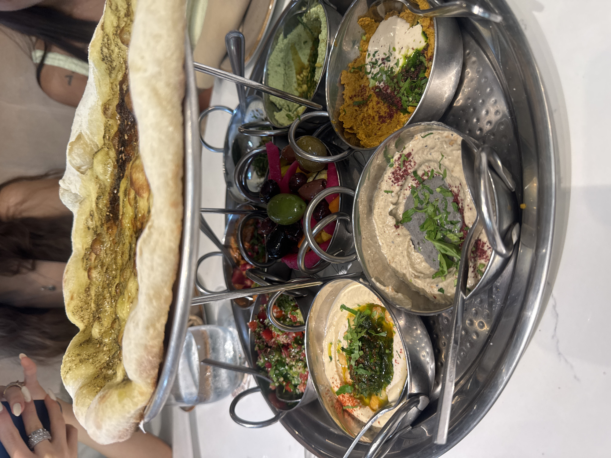 An overhead shot shows a platter of Middle Eastern appetizers at a table in Aventura, Florida. The platter is made of stainless steel and features a raised section in the center, holding a flatbread topped with herbs and oil. Arranged around the central section are several small metal bowls filled with various dips and sides. These include hummus with a drizzle of green sauce and red pepper flakes, a creamy dip topped with herbs and red spice, a yellow dip with a swirl of white sauce and a sprinkle of herbs, a green dip, a salad with chopped tomatoes and parsley, and a selection of olives and pickled vegetables. The platter is placed on a white marble-patterned table. In the background, two women are partially visible, with one in the foreground holding a dark object, possibly a phone. A glass of water is visible to the left of the platter.