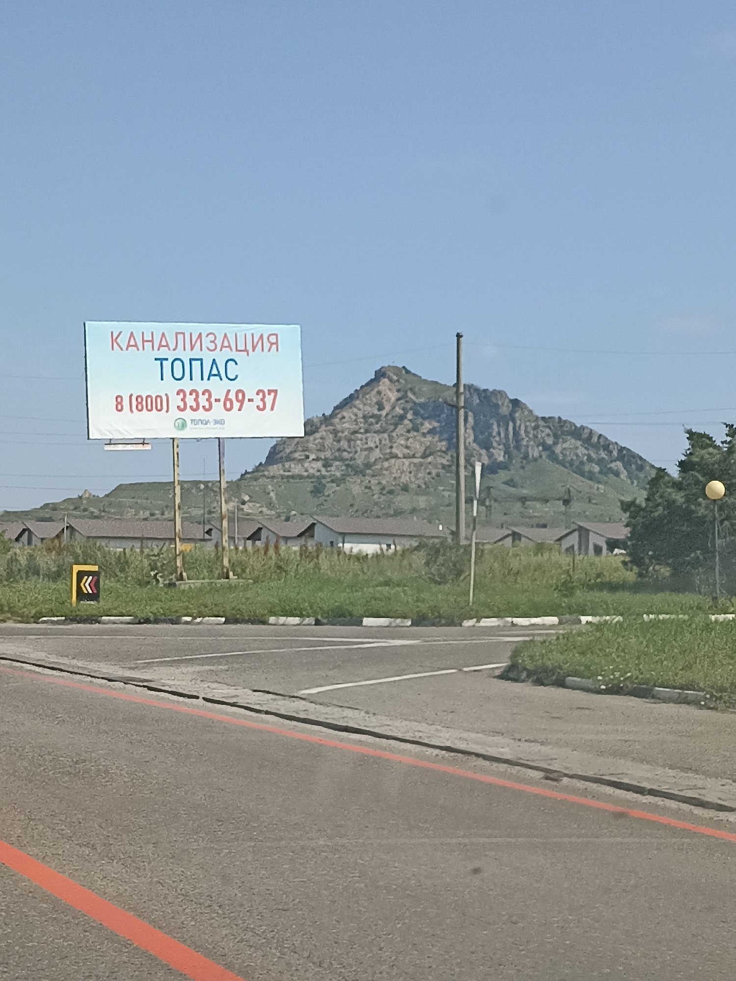 A roadside billboard advertising "KANALIZATSIA TOPAS" with a phone number "8 (800) 333-69-37" is positioned in front of a residential area with numerous houses. A steep, rocky mountain looms in the background. The billboard is mounted on two poles. In the foreground, a road with orange and white markings is visible, along with green grass and shrubs. The sky is clear and blue. The scene appears to be in Vinsady, Russia, based on the provided context.