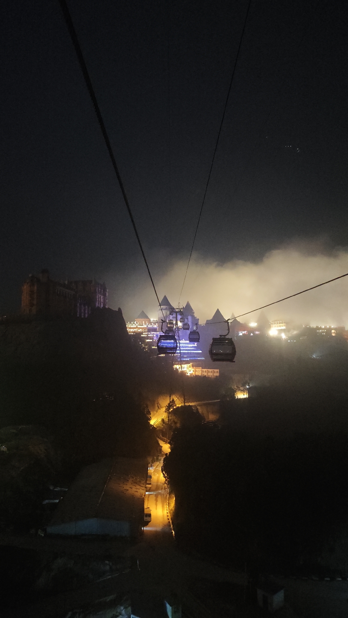The image presents a striking nighttime view from a cable car in Đà Nẵng, Vietnam, likely capturing the scenic Ba Na Hills resort. The dominant subjects are the cable car system itself and an elaborate collection of illuminated buildings on distant hilltops, partially obscured by fog.
In the foreground and upper portions, dark cable car lines stretch diagonally across the frame against a dark, cloudy night sky. A few faint stars are barely visible in the upper right corner. Several cable car gondolas are in motion, with two prominent ones in the middle distance, glowing with a distinct purple and blue internal light.
The scene’s central focus is the architectural landscape. To the left, a large, dark, multi-story building resembling a European castle sits atop a rugged hill. Across a valley, a cluster of ornate buildings, characterized by their peaked roofs and intricate designs, are brightly illuminated. These structures emit a blend of white, blue, purple, and warm orange lights. A thick layer of fog or low clouds blankets the area around these buildings, catching and diffusing the light, which creates an ethereal, glowing effect that lends a fantastical quality to the setting.
In the lower part of the frame, a winding road is visible, illuminated by bright yellow streetlights that carve a path through the dark terrain. Some dark, undefined structures line this road and the surrounding hillsides. The overall impression is a magical, almost otherworldly journey through a mountain resort at night, characterized by vibrant lights piercing through the misty conditions. No people or visible text are discernible.