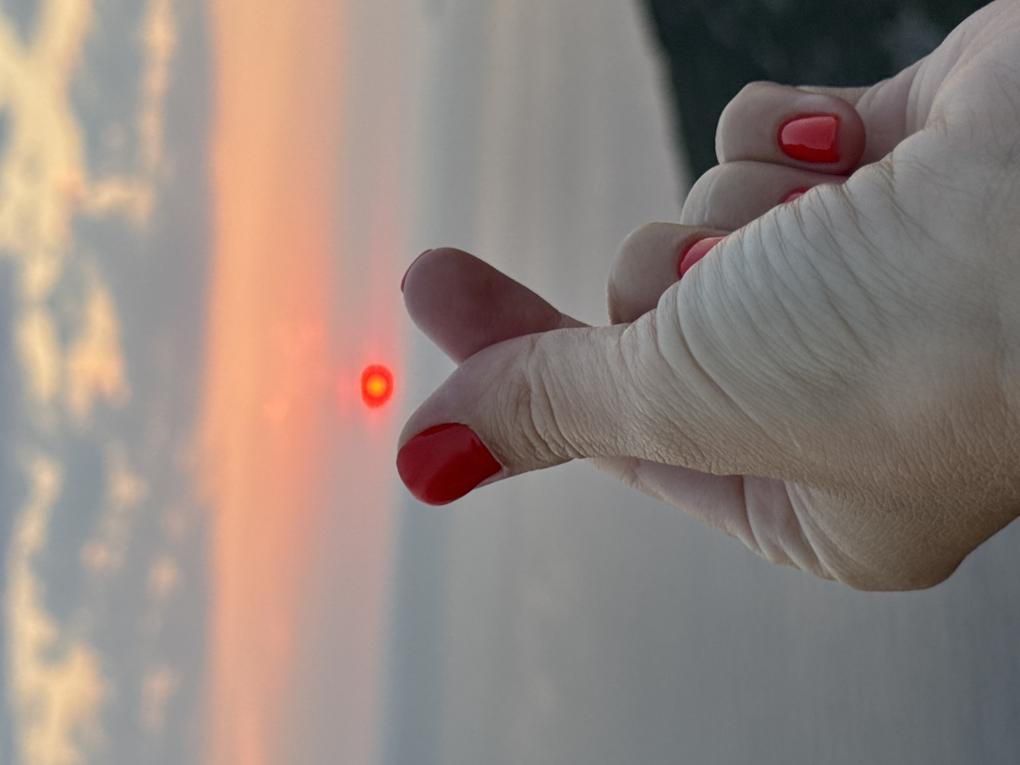An out-of-focus close-up of a hand with red nail polish forms a Korean finger heart gesture in front of a sunset. The sun, a bright red orb, is positioned in the center of the frame, appearing to be held by the hand's gesture. The background consists of a hazy, orange-pink sky with subtle clouds and a calm body of water, likely the Adriatic Sea, stretching to the horizon. The faint outline of a dark, mountainous terrain is visible in the lower right corner, suggesting the location is near the coast of Zagora, Montenegro.