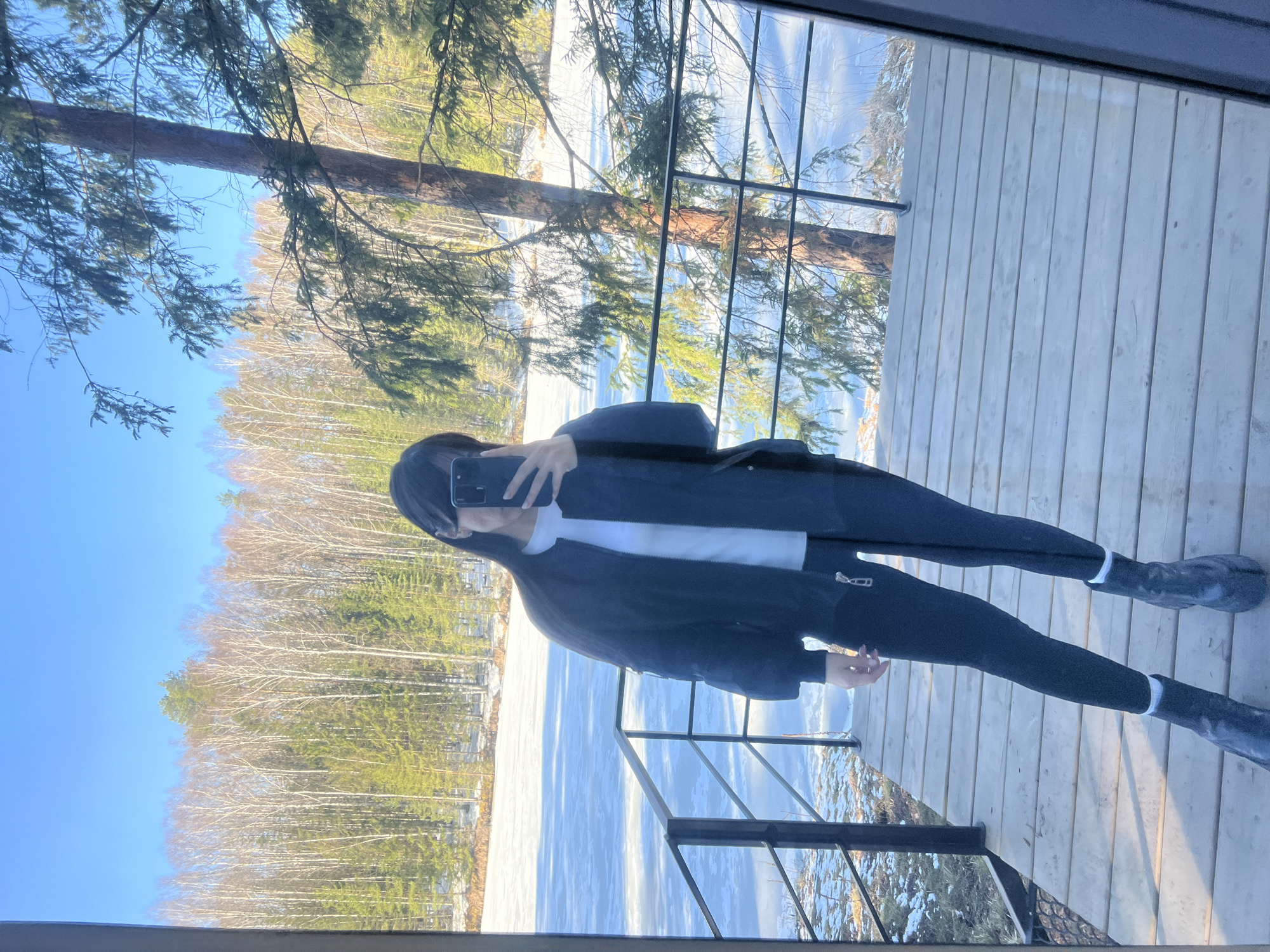A person wearing a black jacket, white top, black pants, and black shoes is taking a picture with their phone in a mirror. The reflection shows a winter landscape with a frozen lake or river surrounded by trees. The background consists of a forest of deciduous trees without leaves, along with some evergreen trees. The sky is blue, indicating a clear day. The person is standing on a wooden deck, as can be seen in the mirror. The location is Мишкино, Russia.