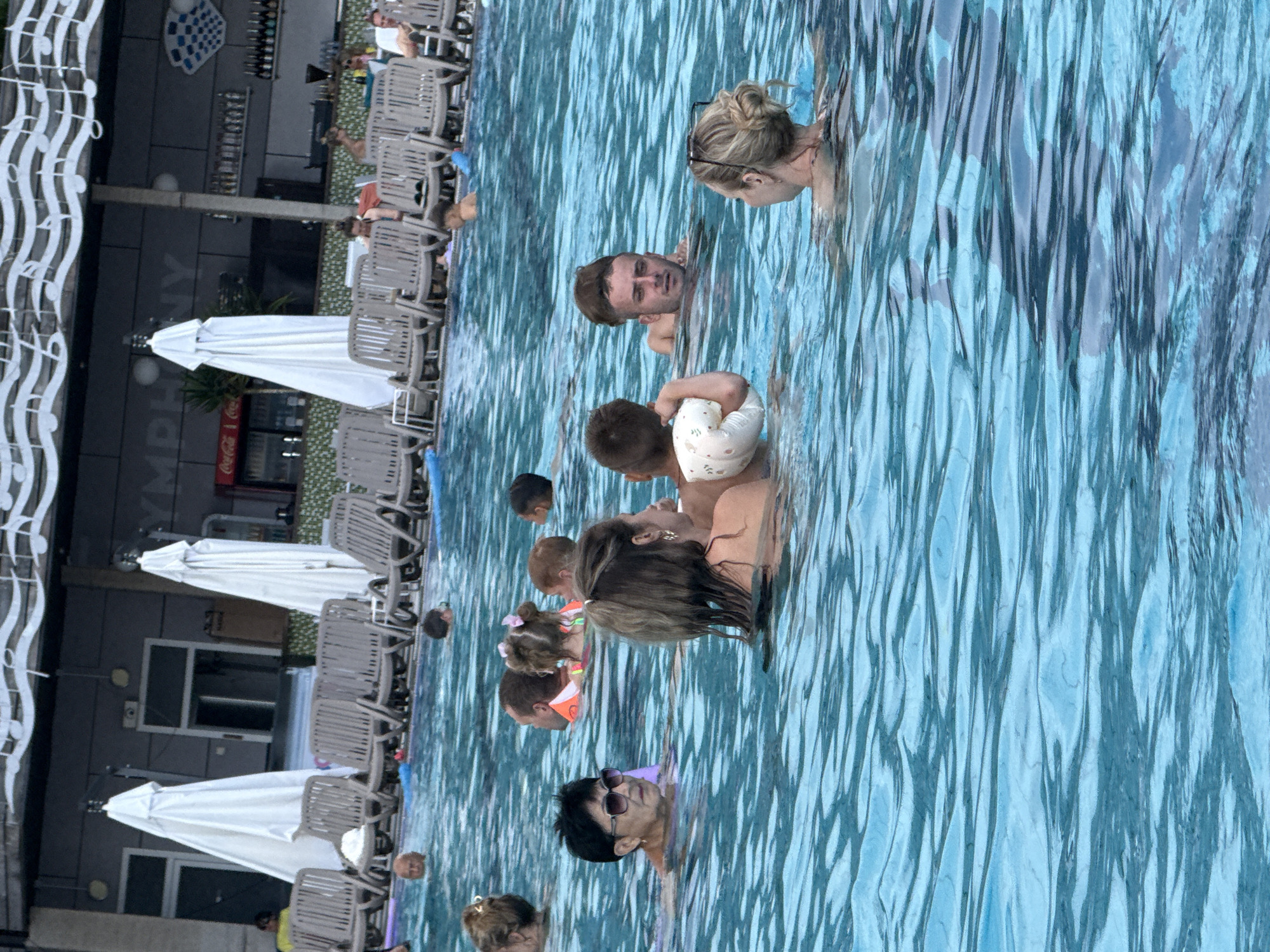 The image depicts a crowded swimming pool scene, likely at a resort or public facility. In the foreground, several people are visible in the water, some swimming and others wading. A woman with sunglasses and short dark hair is in the lower left of the frame. To her right, a woman with her back to the camera and blonde hair pulled up is holding a young child. Another man with short, dark hair is swimming behind them. In the middle ground, a group of people are in the water, including a child with a pink bow in her hair and an adult with an orange floatation device. Several lounge chairs are visible behind the pool, along with white umbrellas. A building with a sign that appears to read "SYMPHONY" is in the background, along with a bar area and refrigerators. The water is a clear blue, with ripples on the surface indicating movement. The overall atmosphere suggests a lively and recreational setting.