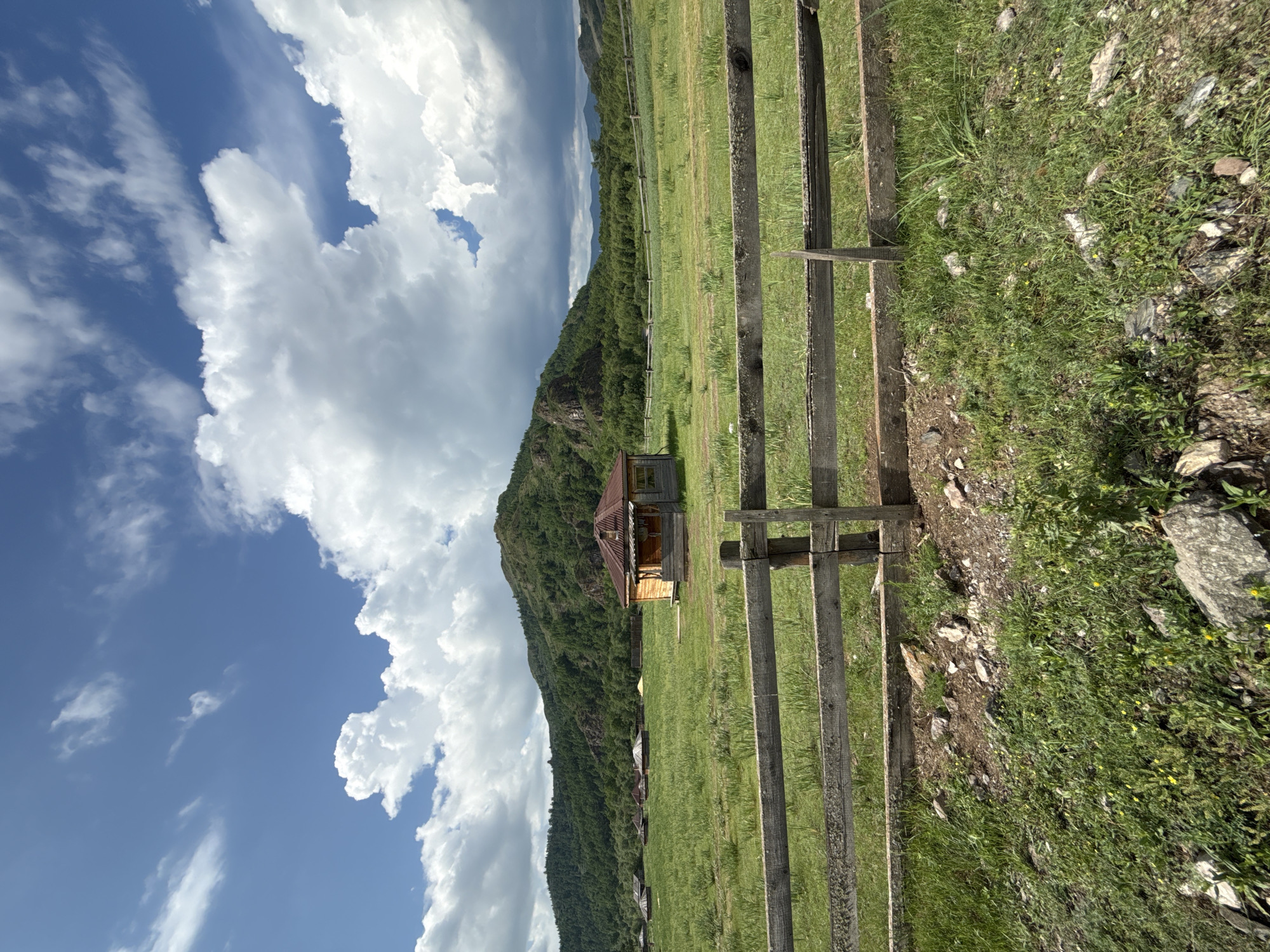 The image depicts a rural landscape with a traditional wooden house and several yurts in a grassy field. A rustic wooden fence runs across the foreground. In the background, a verdant, tree-covered mountain rises under a dramatic sky with large, white clouds. The scene is set in the village of Kazanovka, Russia, in a natural environment.