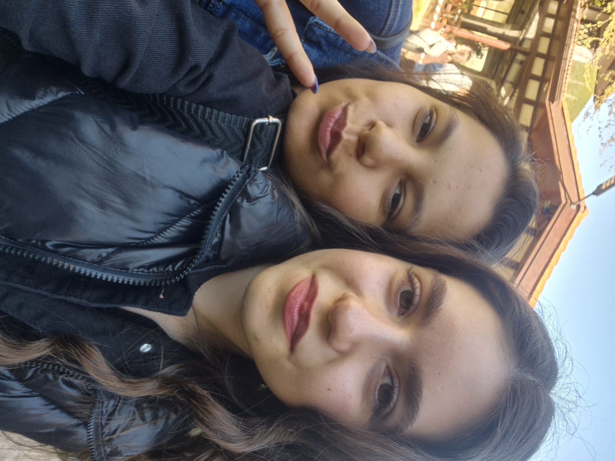 An outdoor portrait captures two young women posing for a selfie, rotated about 90 degrees to the right. Both women are shown from the chest up, positioned closely together and looking towards the camera with soft smiles or smirks.
The woman in the foreground has long, wavy dark brown hair, striking brown eyes, and is wearing red lipstick. She is dressed in a black, shiny puffer jacket with visible zipper details and a silver buckle on a strap across her chest. Her expression is calm and pleasant.
Behind her, the second woman has dark brown hair and brown eyes. She is wearing a dark, possibly black, jacket layered with a blue denim jacket. Her right hand is raised, making a peace sign with painted fingernails, indicating a playful or friendly mood.
The background reveals a sunny outdoor setting with a clear blue sky. Parts of a building structure are visible, featuring light-colored or wooden beams and roof sections, along with hints of green and yellow foliage, suggesting a natural or rustic environment. The lighting is bright and even, consistent with daytime. No visible text is present in the image. The scene is set in Devin, Bulgaria.