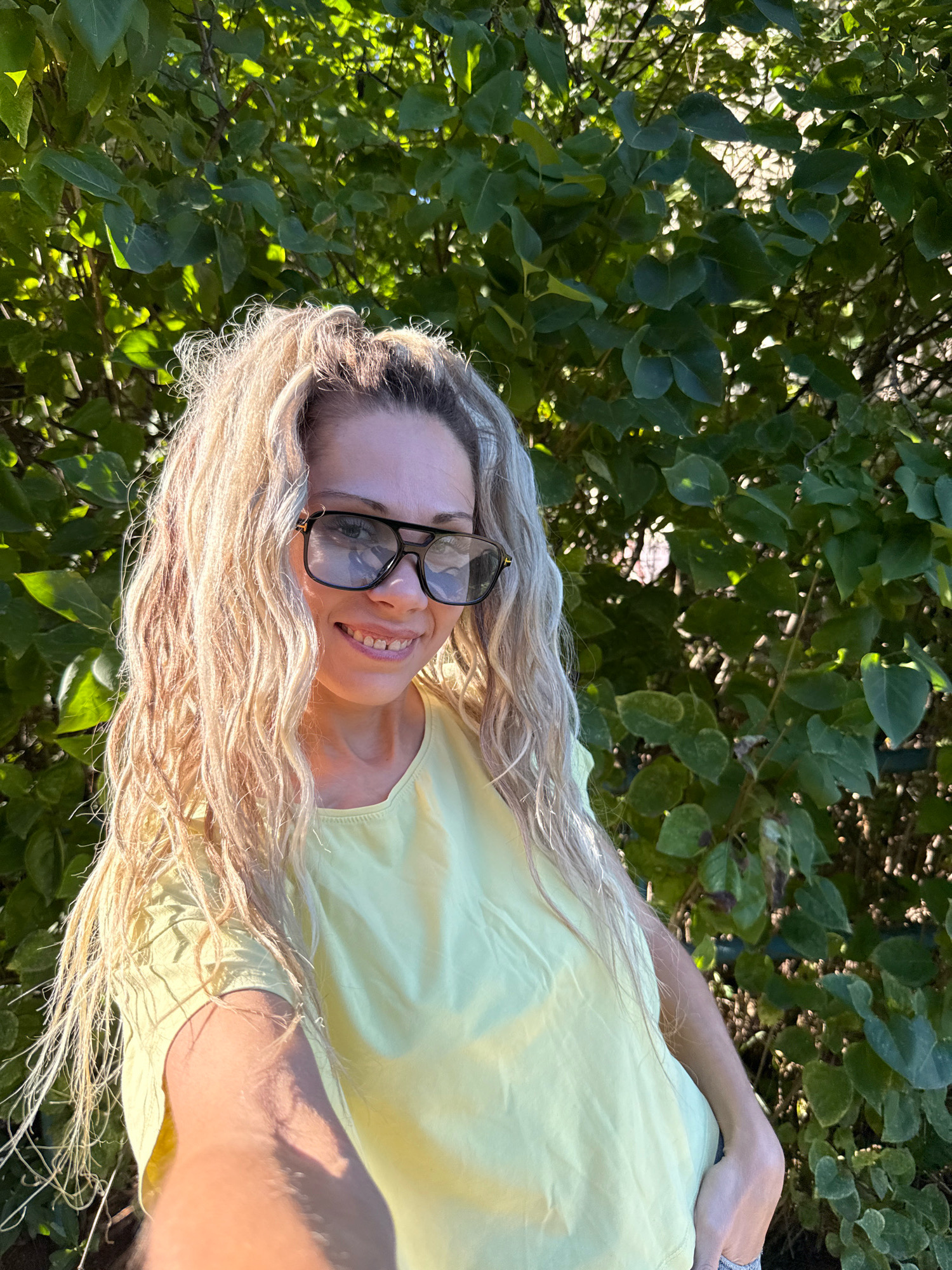 In the city of Rusanovo, Russia, a woman with long, wavy blonde hair is captured in a selfie taken outdoors. She is wearing large, dark-rimmed glasses and a light yellow top. The background is filled with lush green foliage, suggesting a natural or garden setting. The lighting indicates it is daytime, with sunlight filtering through the leaves. The woman is smiling and looking at the camera, with her arm extended to take the photo.