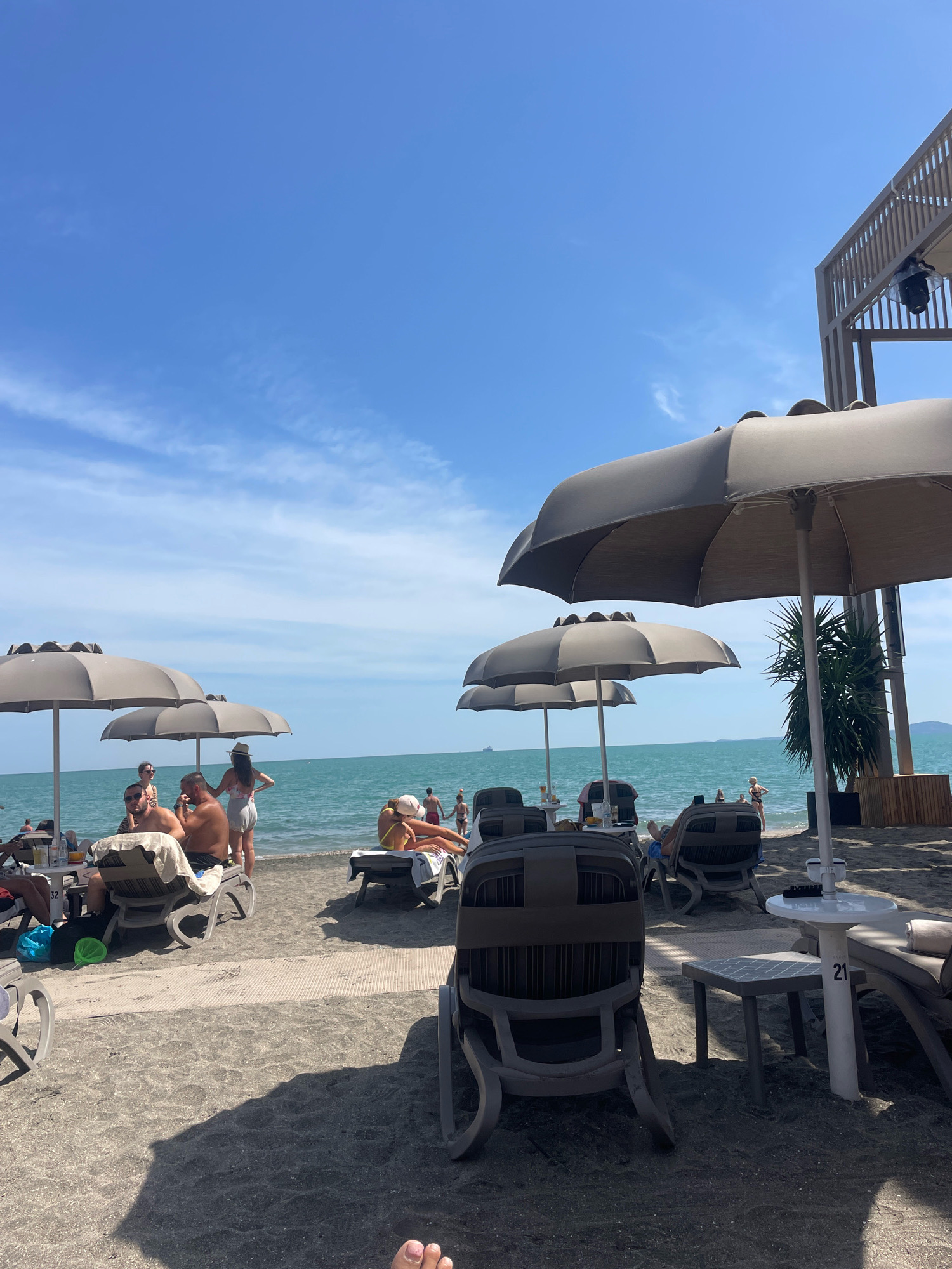 The image depicts a sunny day at a beach resort, likely in Burgas, Bulgaria, given the context of the request. The scene is focused on a stretch of dark sand leading to the turquoise waters of the sea. Several sun loungers are arranged on the beach, some occupied by people relaxing or socializing. Large, neutral-colored umbrellas provide shade over some of the loungers. In the foreground, a plastic sun lounger and a small side table with the number "21" are visible. The sea is calm, and a few individuals are seen wading or standing near the water's edge. In the background, there are more beachgoers, umbrellas, and a structure that appears to be part of the resort, possibly a bar or restaurant with wooden accents and an awning. The sky is bright blue with scattered white clouds. A few drinks and a plastic bag are placed on a table near some of the loungers.