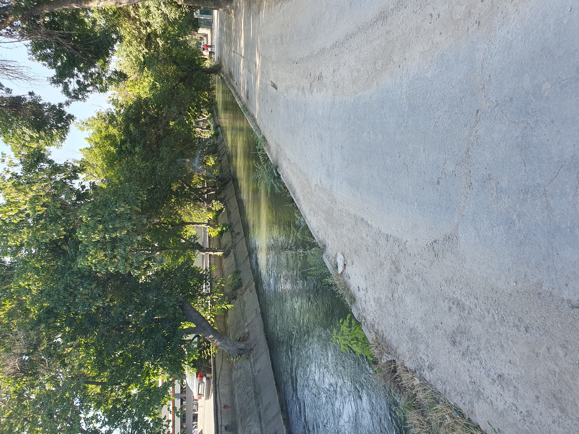 The image shows a straight, concrete-lined canal with flowing water. A paved pathway runs alongside the canal, bordered by trees. In the distance, two people are walking away from the camera on the pathway. On the left side of the canal, there is a road with a parked car. The scene appears to be a public area within a city, likely Tashkent, Uzbekistan, given the characteristic architecture of the canal and the presence of trees commonly found in the region. The lighting suggests it is daytime, with sunlight casting shadows. The overall atmosphere is calm and serene, depicting a typical urban landscape with green spaces.
