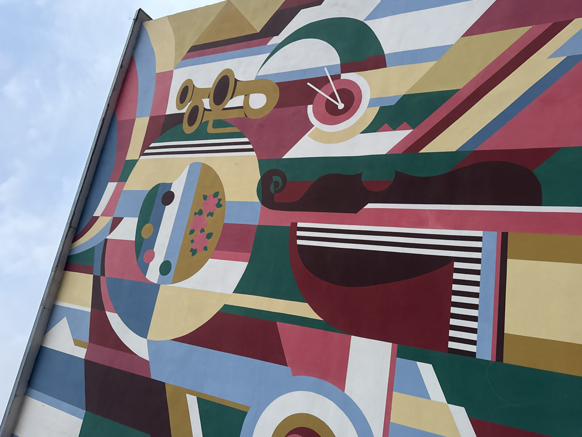 This is a photograph of a mural on the exterior wall of a building in Urzhum, Russia. The mural is an abstract geometric design with bold colors including blues, reds, greens, yellows, and whites. The artwork features various shapes such as circles, squares, rectangles, and curved lines, arranged in a dynamic composition. Some sections of the mural appear to depict stylized musical instruments, including what looks like a trumpet and a cello. The mural covers a significant portion of the building's facade, extending upwards towards the sky. The sky is visible in the upper right corner of the image, with some clouds present. The building itself appears to be a multi-story structure, with a concrete or metal gutter visible along the top edge of the wall. No people are visible in the image. The overall impression is one of vibrant, modern public art.